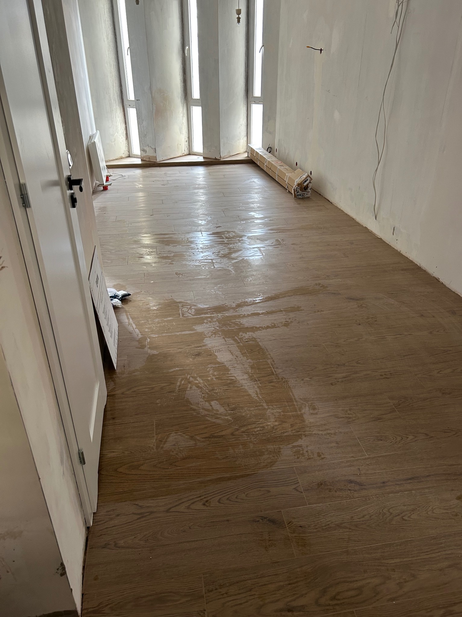 Ułożona podłoga z płytek imitujących drewno w jasnym pomieszczeniu w trakcie prac wykończeniowych. Widoczne okna, drzwi i materiały budowlane.