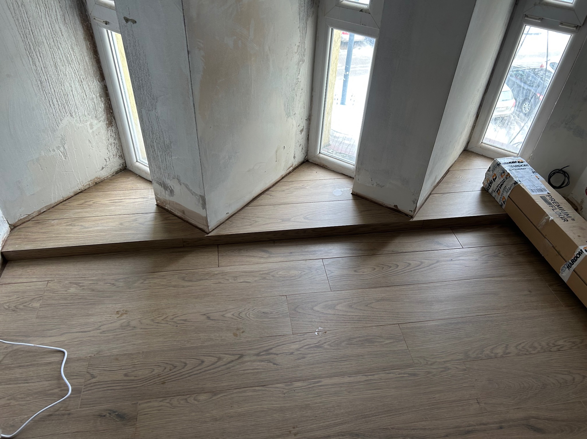 Układanie paneli podłogowych w nietypowym pomieszczeniu z oknami wnękowymi. Widoczne panele i fragment ściany przed wykończeniem. Częściowo ułożona podłoga.