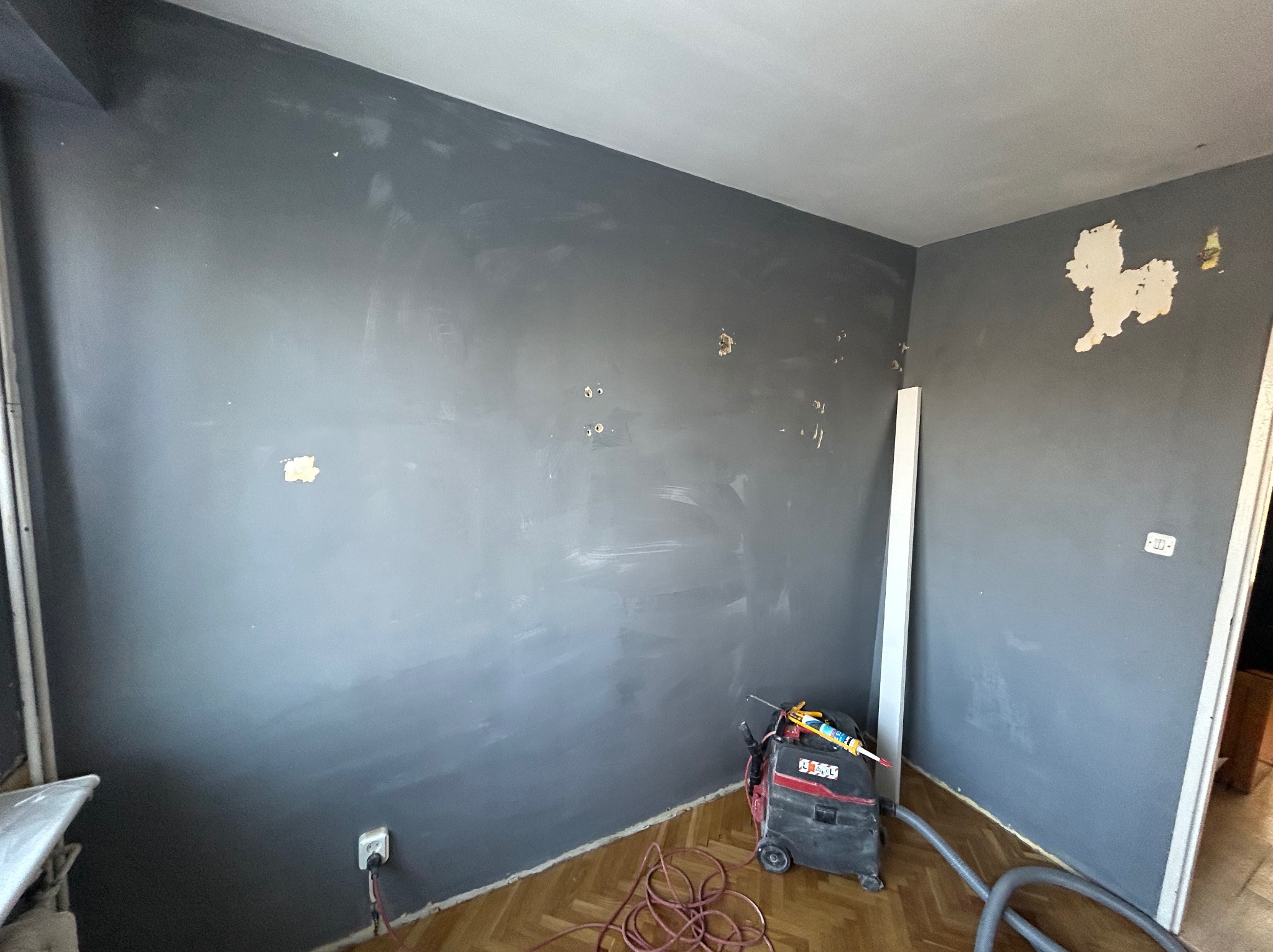 Pokój w trakcie przygotowania do malowania: szare ściany z widocznymi śladami po szpachlowaniu, odkurzacz przemysłowy i parkiet na podłodze. Widoczne gniazdko elektryczne.