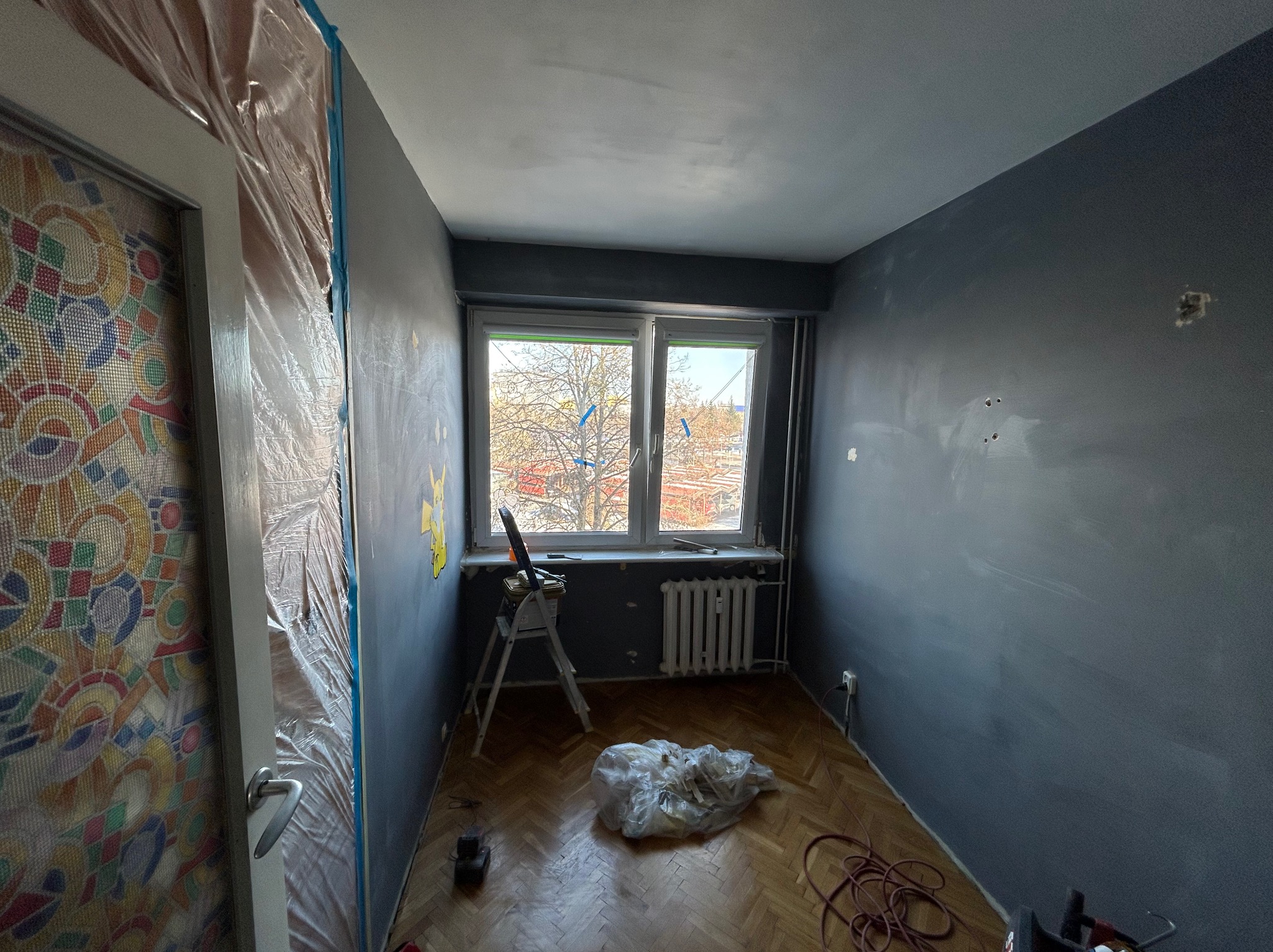 Pokój w trakcie malowania na szaro, zabezpieczone drzwi folią, drabina przy oknie, widoczne ślady szpachlowania na ścianach, parkiet na podłodze.