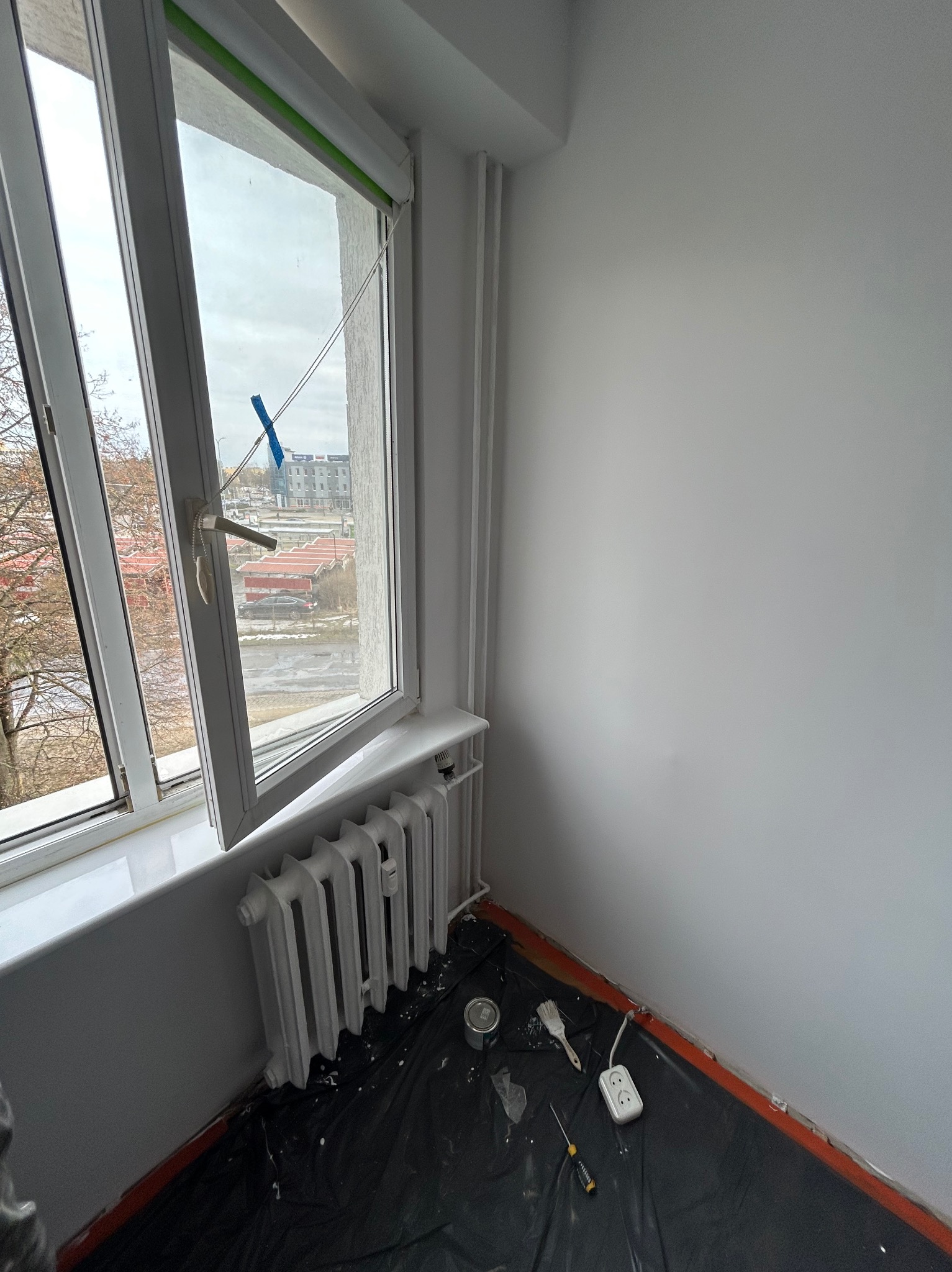 Pokój w trakcie malowania: zabezpieczona podłoga, otwarte okno z widokiem na zewnątrz, pomalowany grzejnik i ściana. Widoczne narzędzia malarskie.