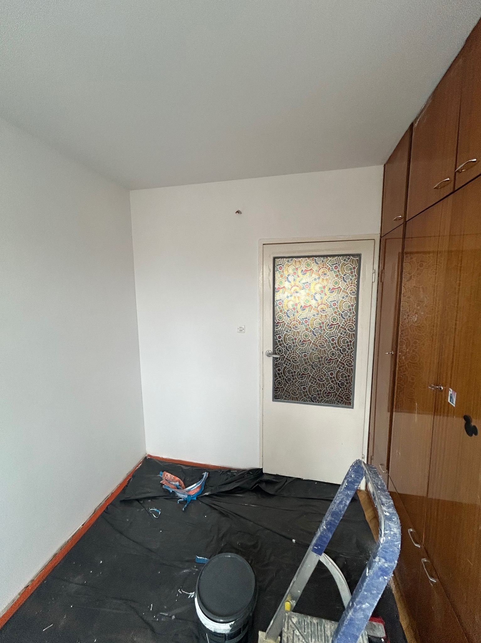 Pokój w trakcie malowania: świeżo pomalowane białe ściany, zabezpieczona podłoga czarną folią, drabina i puszka z farbą. Drzwi z ozdobnym szkłem i szafa w tle.