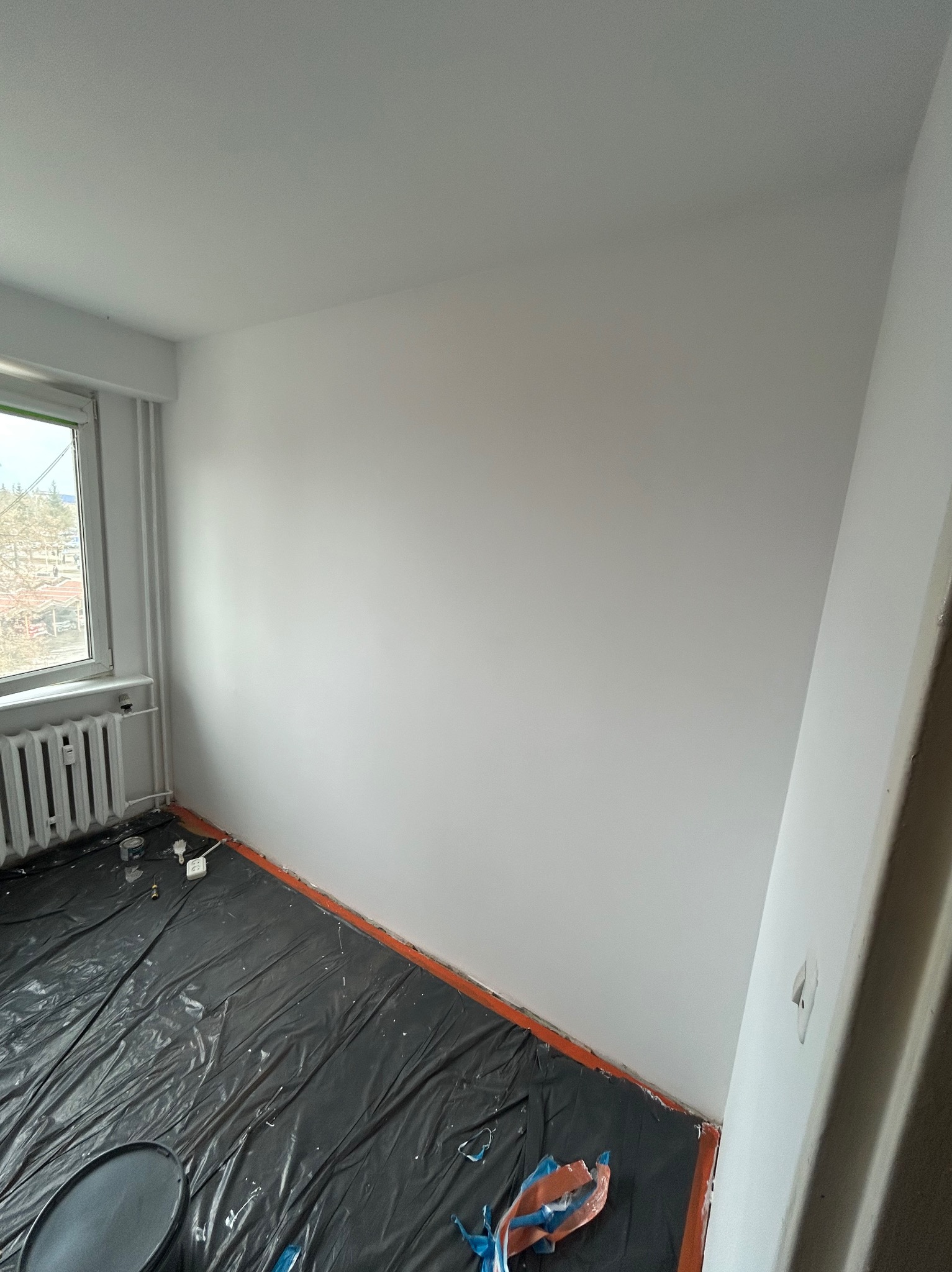 Świeżo pomalowane na biało wnętrze pokoju z zabezpieczoną folią podłogą. Widoczne okno z grzejnikiem i fragment drzwi. Minimalistyczny kadr, jasne oświetlenie.