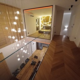 GALSAN SPÓŁKA Z OGRANICZONĄ ODPOWIEDZIALNOŚCIĄ - Nowoczesne wnętrze domu z drewnianą podłogą w jodełkę, szklaną balustradą i wiszącą lampą z kulistymi kloszami. Widoczny fragment sypialni z oświetleniem LED na ścianie.