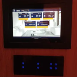 Panel sterowania inteligentnym domem z ekranem dotykowym pokazującym temperatury w różnych pomieszczeniach i wyłączone funkcje, zamontowany na ścianie o fakturze drewna, poniżej panel dotykowy...