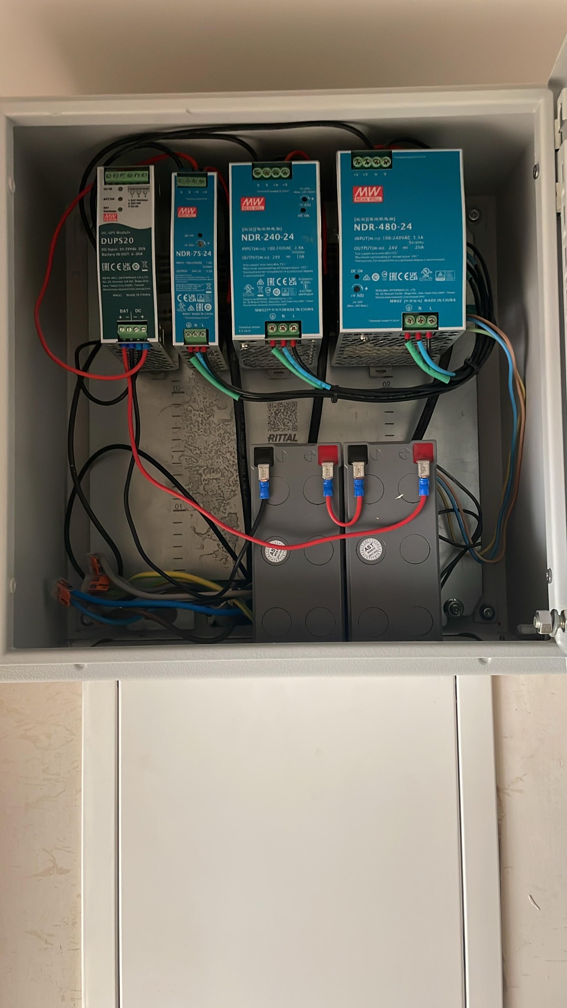 Wnętrze skrzynki elektrycznej z czterema zasilaczami Mean Well (DUPS20, NDR-75-24, NDR-240-24, NDR-480-24) i dwoma akumulatorami AST, uporządkowane okablowanie.