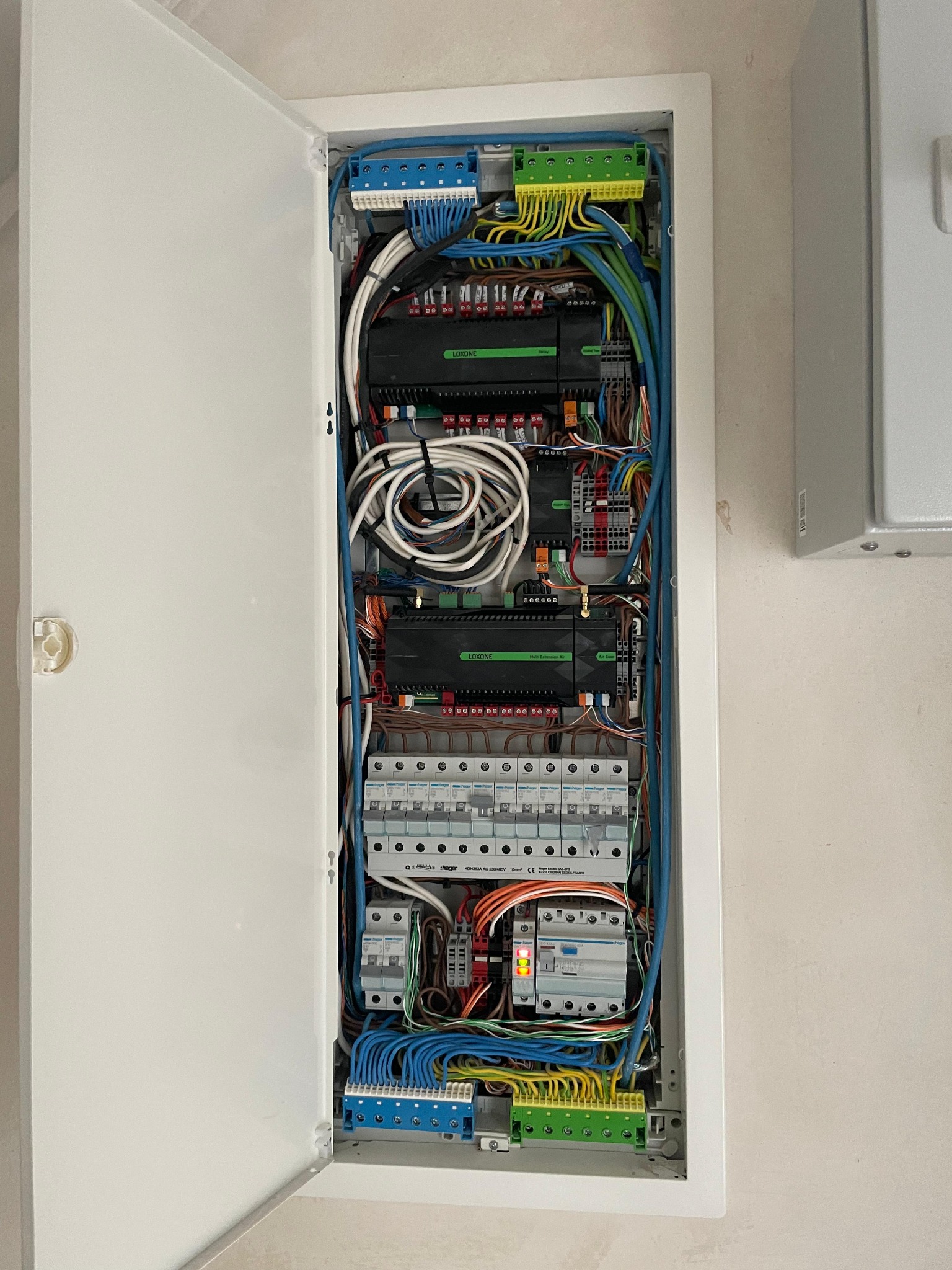 Otwarta szafa rozdzielcza z zainstalowanymi modułami Loxone, bezpiecznikami Hager i licznymi przewodami w kolorach niebieskim, zielonym i białym, zamocowanymi do listew zaciskowych.