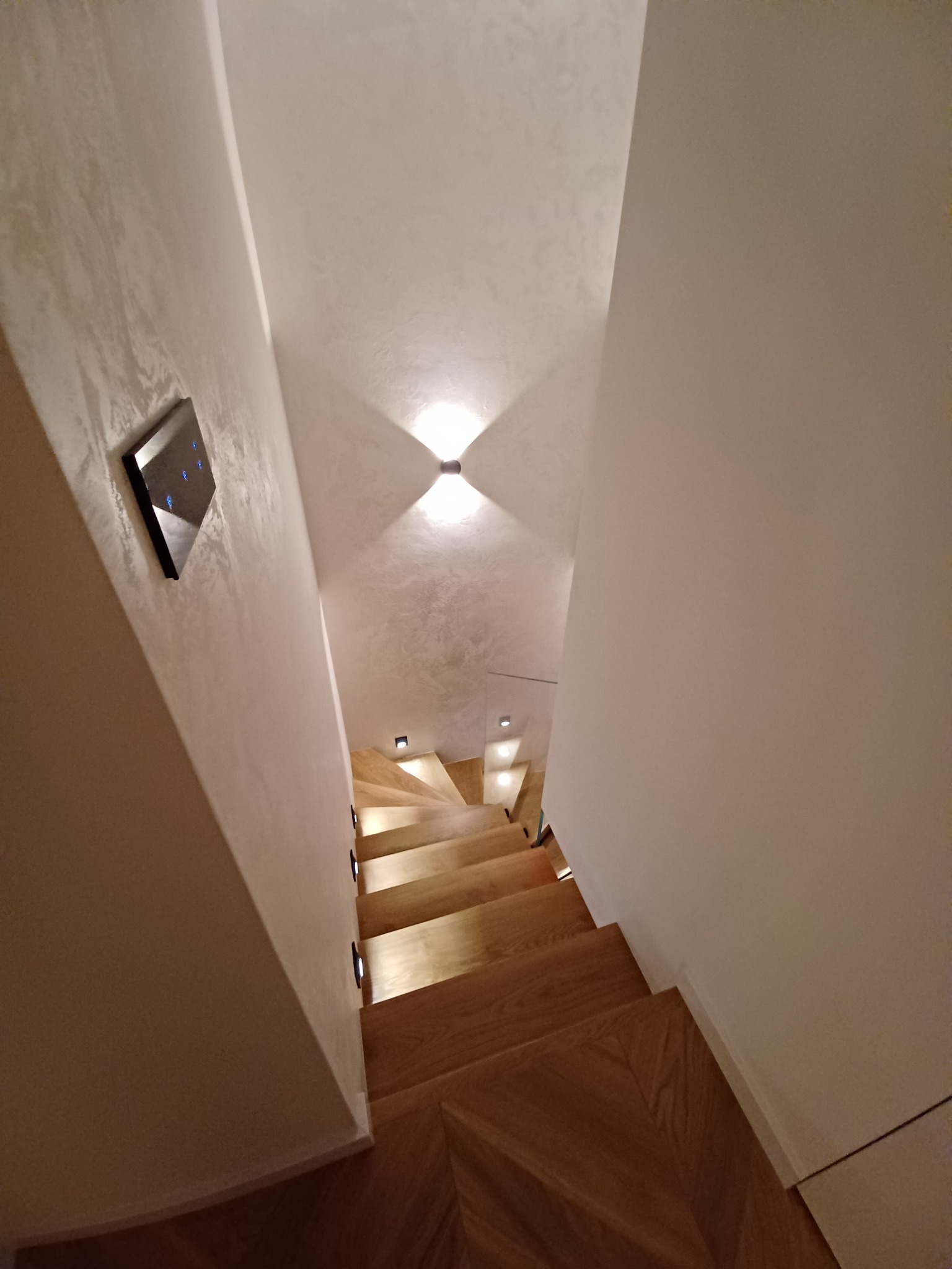 Klatka schodowa z drewnianymi schodami oświetlonymi punktowymi lampami LED w ścianie i na suficie, widoczny panel sterowania inteligentnego domu na ścianie z dekoracyjną fakturą.