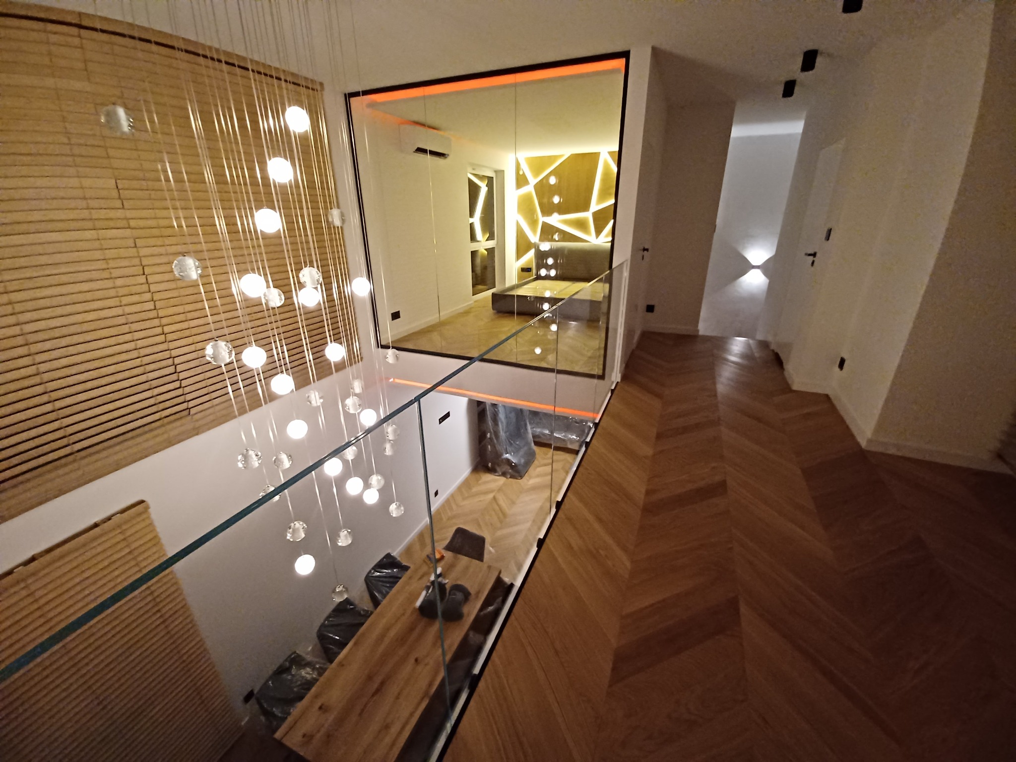 Nowoczesne wnętrze domu z drewnianą podłogą w jodełkę, szklaną balustradą i wiszącą lampą z kulistymi kloszami. Widoczny fragment sypialni z oświetleniem LED na ścianie.