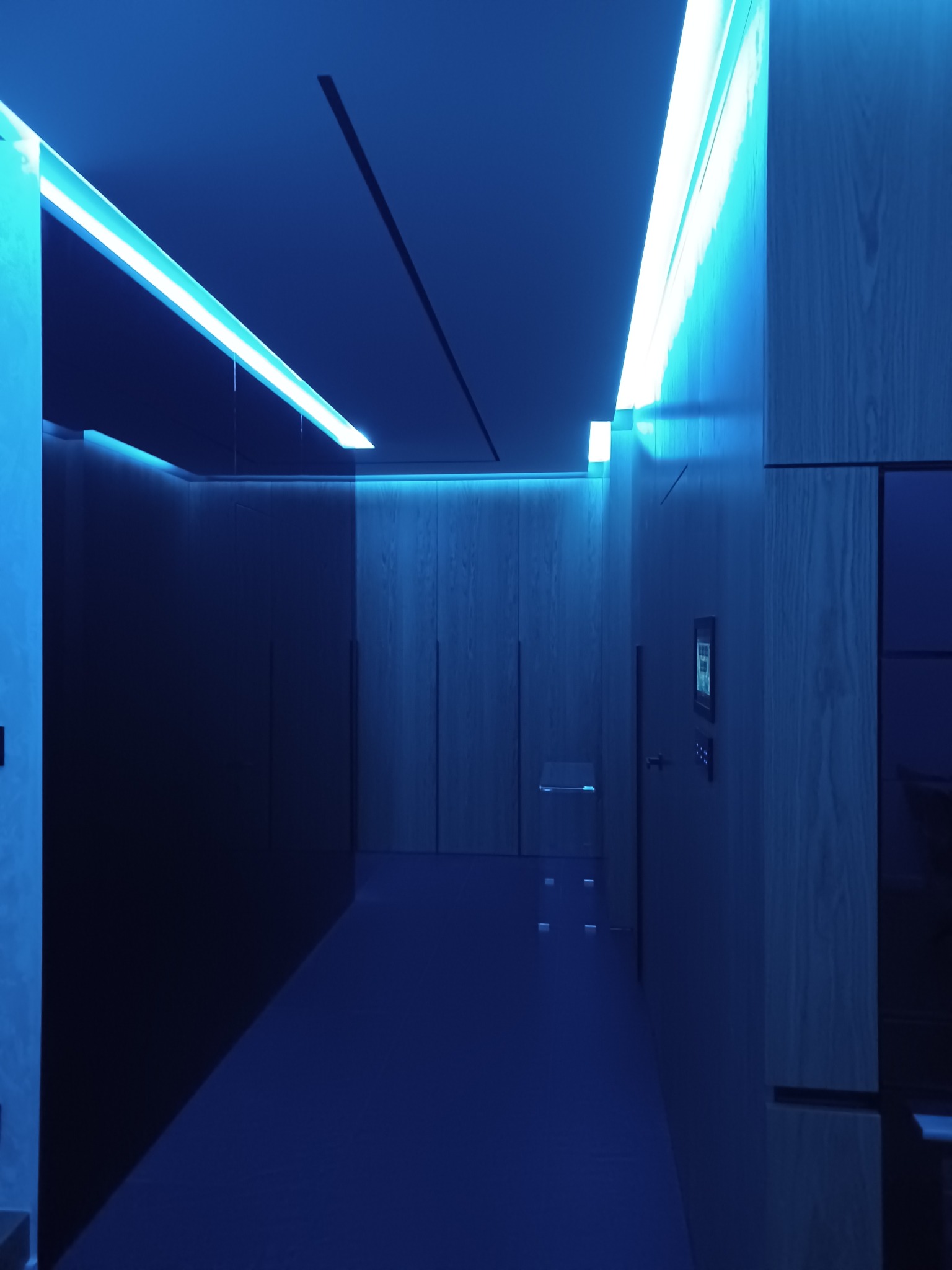 Korytarz z oświetleniem LED w odcieniach niebieskiego, podwieszany sufit z ukrytymi lampami, drewniane panele ścienne, minimalistyczny design.