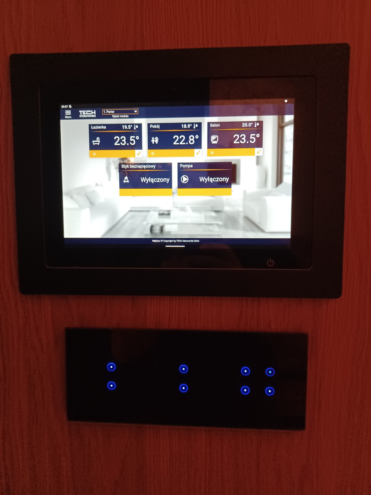 Panel sterowania inteligentnym domem z ekranem dotykowym pokazującym temperatury w różnych pomieszczeniach i wyłączone funkcje, zamontowany na ścianie o fakturze drewna, poniżej panel dotykowy...