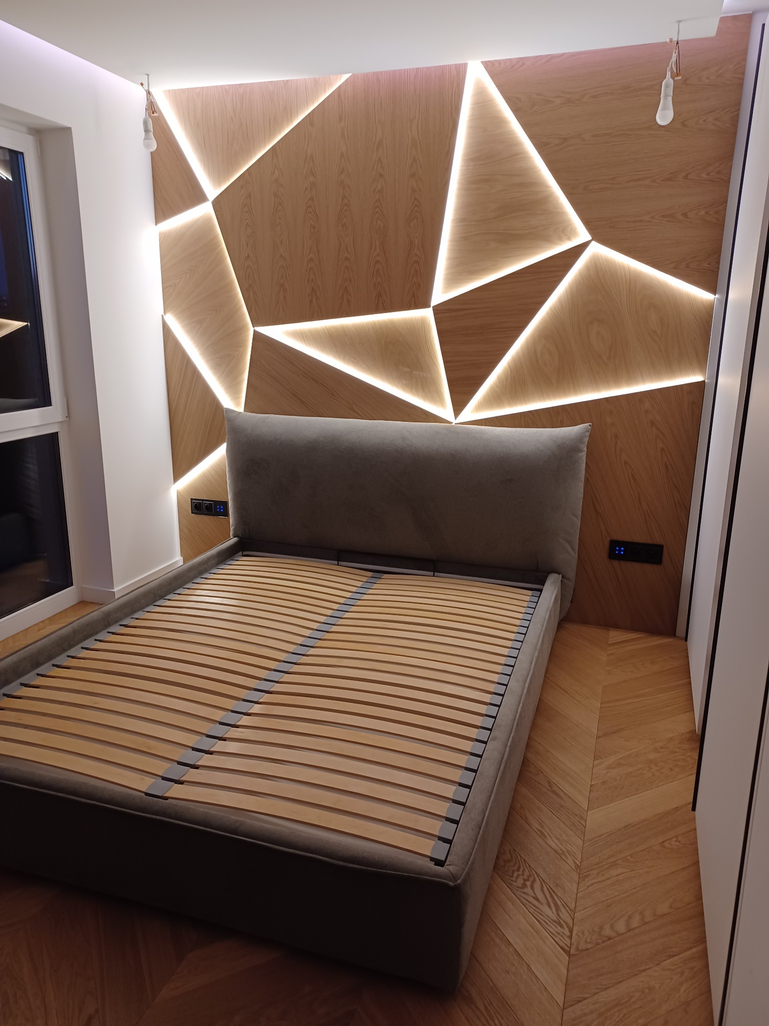 Nowoczesna sypialnia z drewnianym panelem ściennym o geometrycznym wzorze podświetlonym ledami, szare łóżko z drewnianym stelażem i parkiet ułożony w jodełkę.