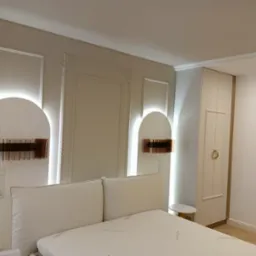 Elegancka sypialnia z beżowymi ścianami, dekoracyjnymi listwami, podświetlanymi wnękami i nowoczesnymi lampkami nocnymi z brązowymi akcentami, widok na łóżko z kremowym zagłówkiem i szafę...