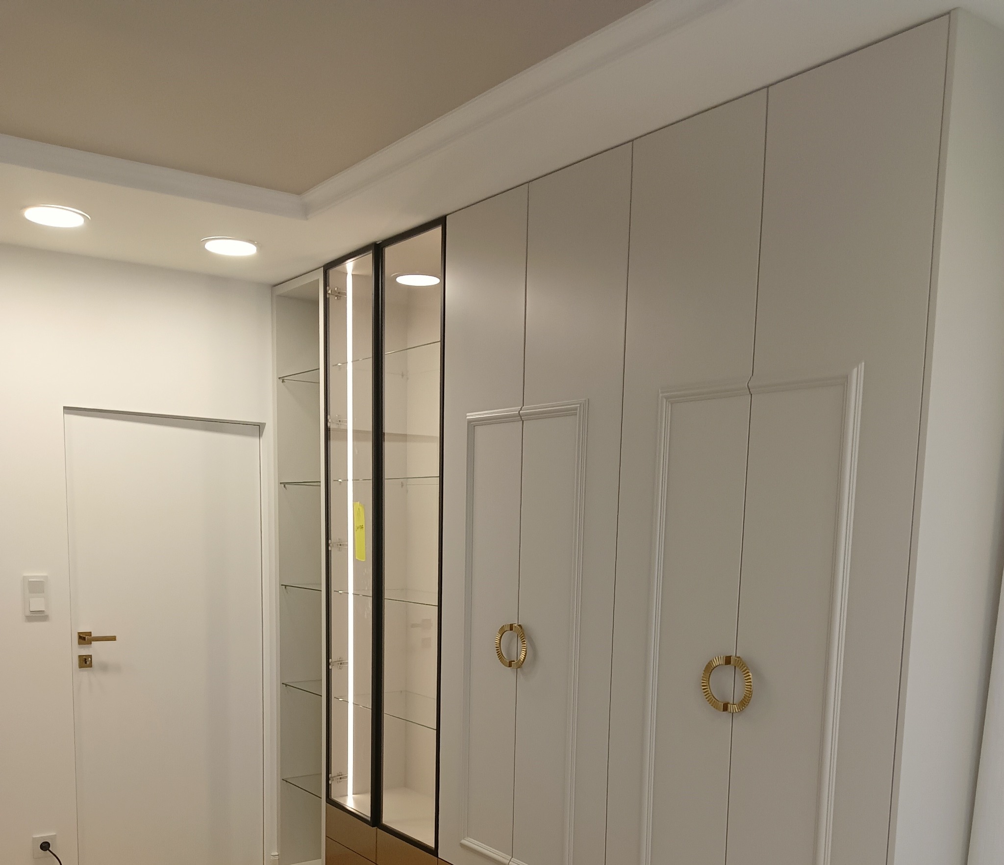 Biała zabudowa meblowa z przeszkloną witryną z oświetleniem LED i dekoracyjnymi, okrągłymi uchwytami na frontach, w minimalistycznym wnętrzu.