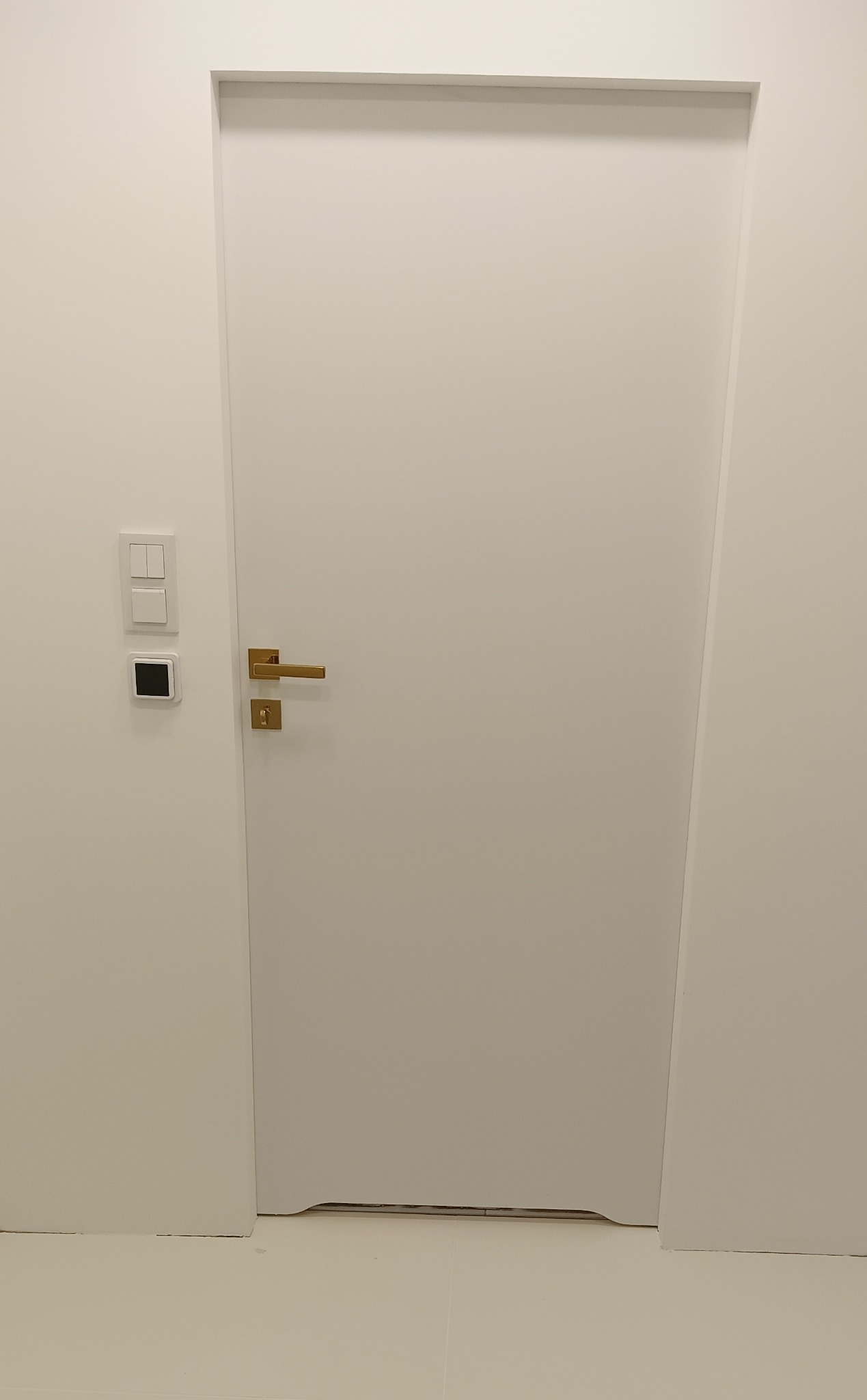 Minimalistyczne, białe drzwi wewnętrzne z złotą klamką w Gorenice, zamontowane w ścianie z widocznymi włącznikami światła i termostatem.