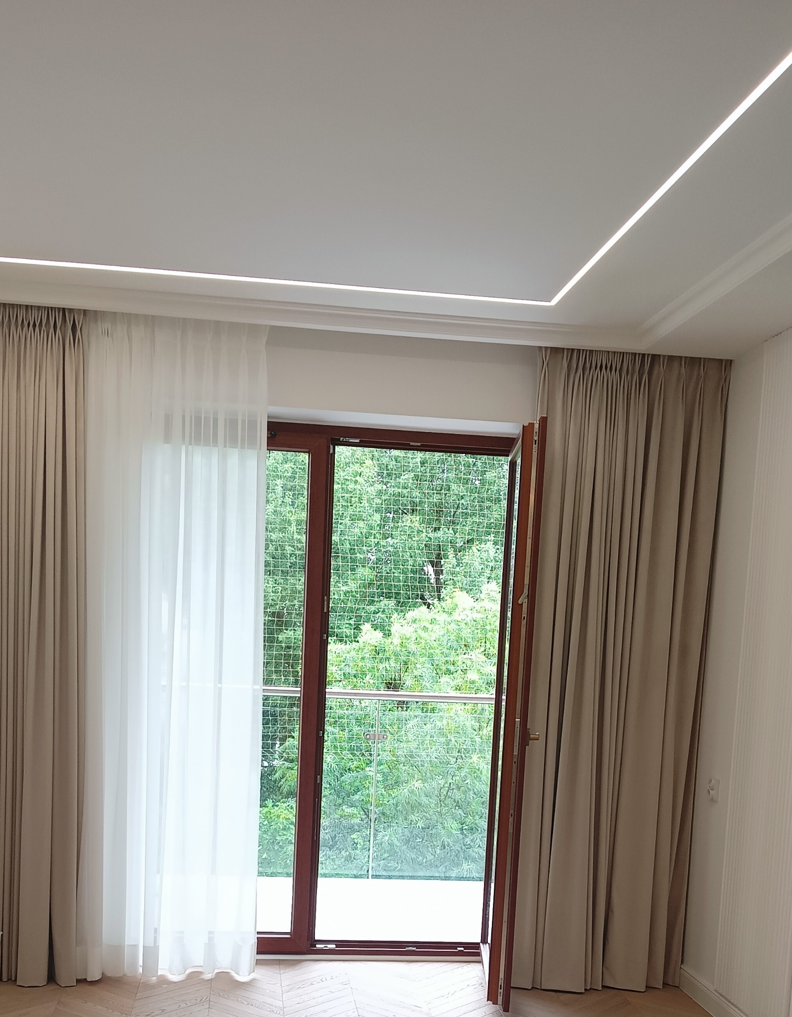 Nowoczesne wnętrze z drewnianą stolarką okienną, beżowymi zasłonami, białymi firanami i subtelnym oświetleniem LED na suficie.