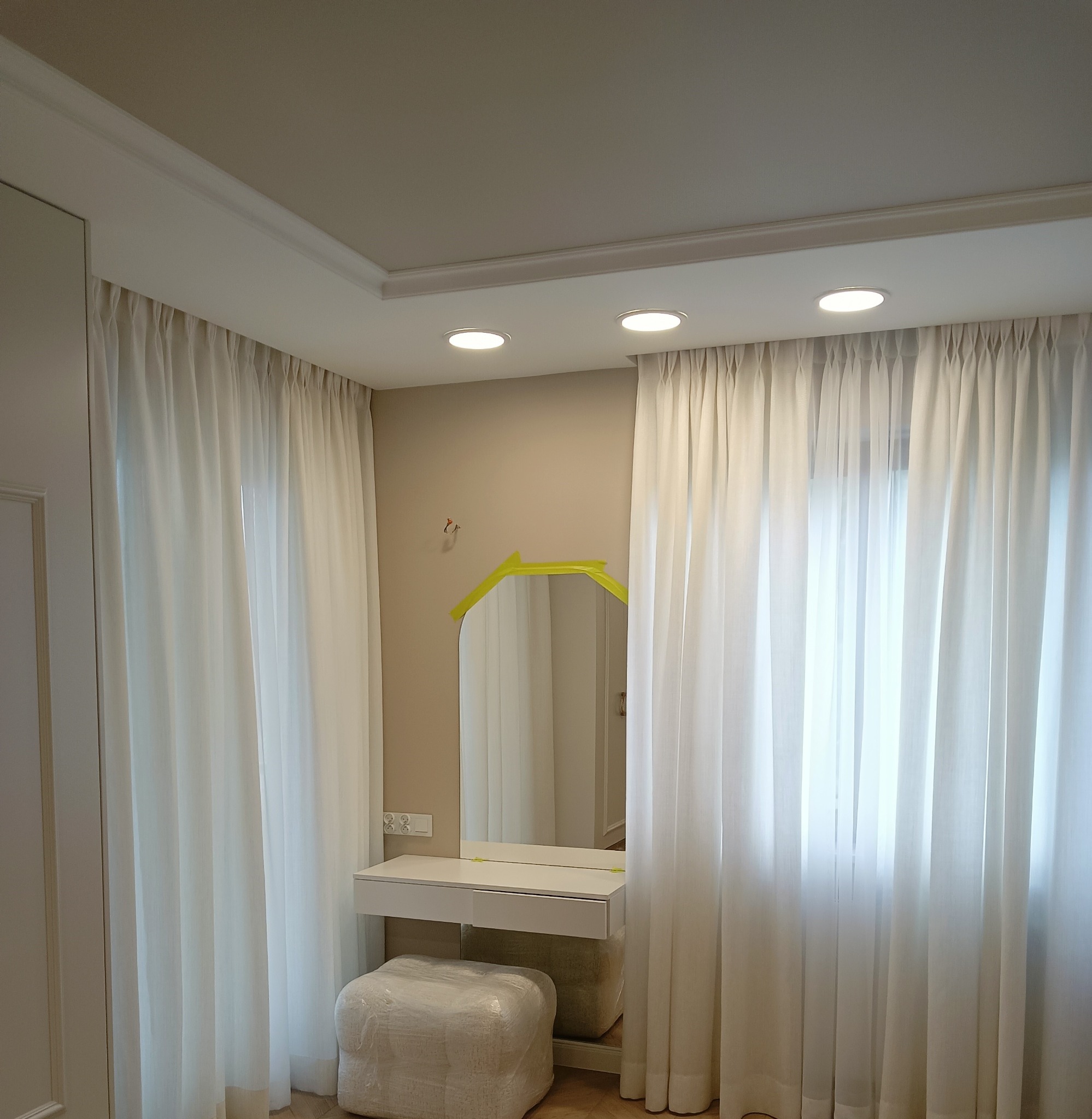 Remont łazienki w trakcie: białe zasłony, toaletka z lustrem zabezpieczonym taśmą malarską, pufa owinięta folią ochronną, sufit z oświetleniem punktowym.