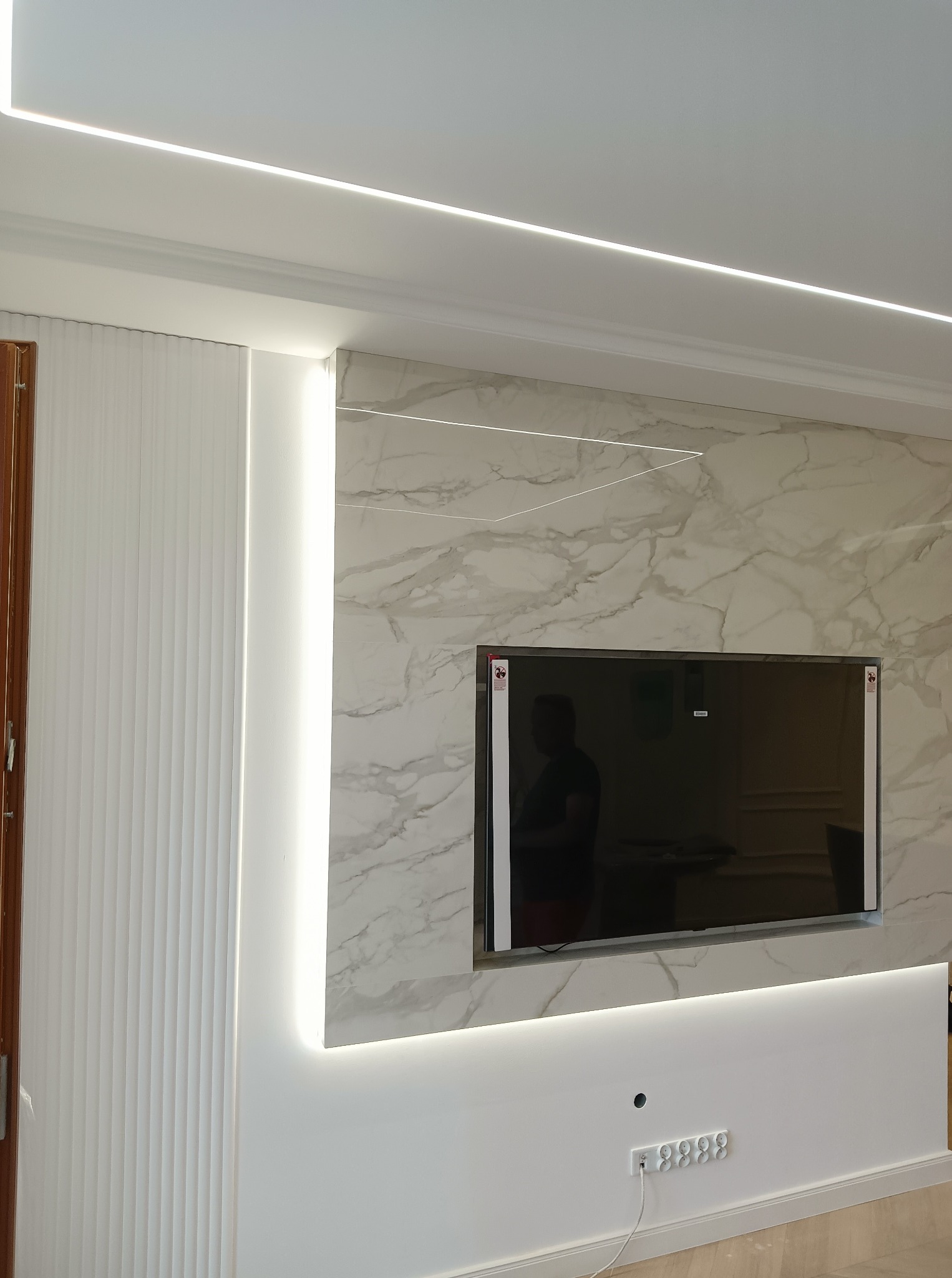 Telewizor zawieszony na ścianie z marmurowym wzorem, podświetlony listwami LED, widoczne gniazdka elektryczne i dekoracyjne panele ścienne z pionowymi żłobieniami.