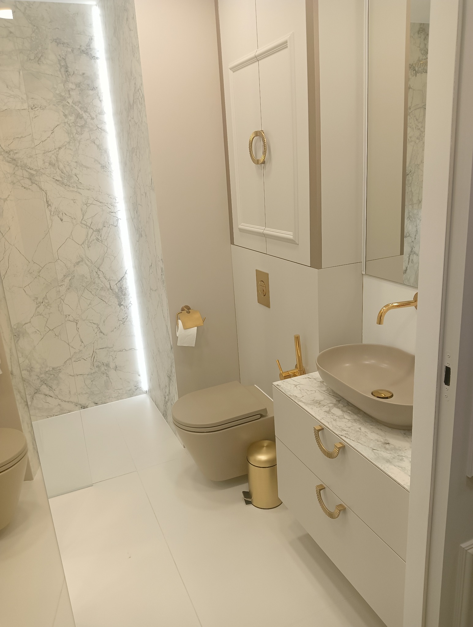 Elegancka łazienka w beżach i bieli z marmurową ścianą podświetloną LED, wisząca toaleta, umywalka nablatowa na szafce z marmurowym blatem i złotymi uchwytami, złote dodatki.