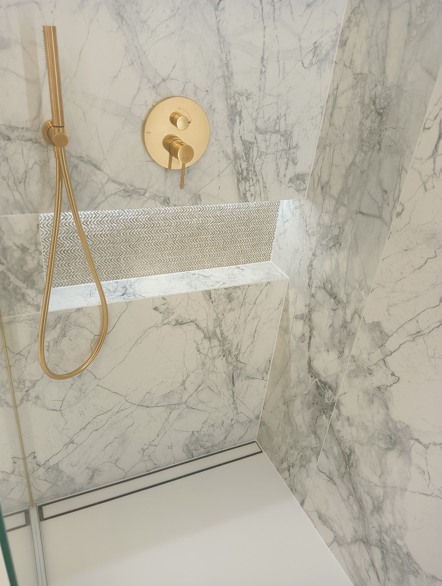 Wykończona kabina prysznicowa z marmurowymi płytami, złotą armaturą i wnęką z mozaiką.