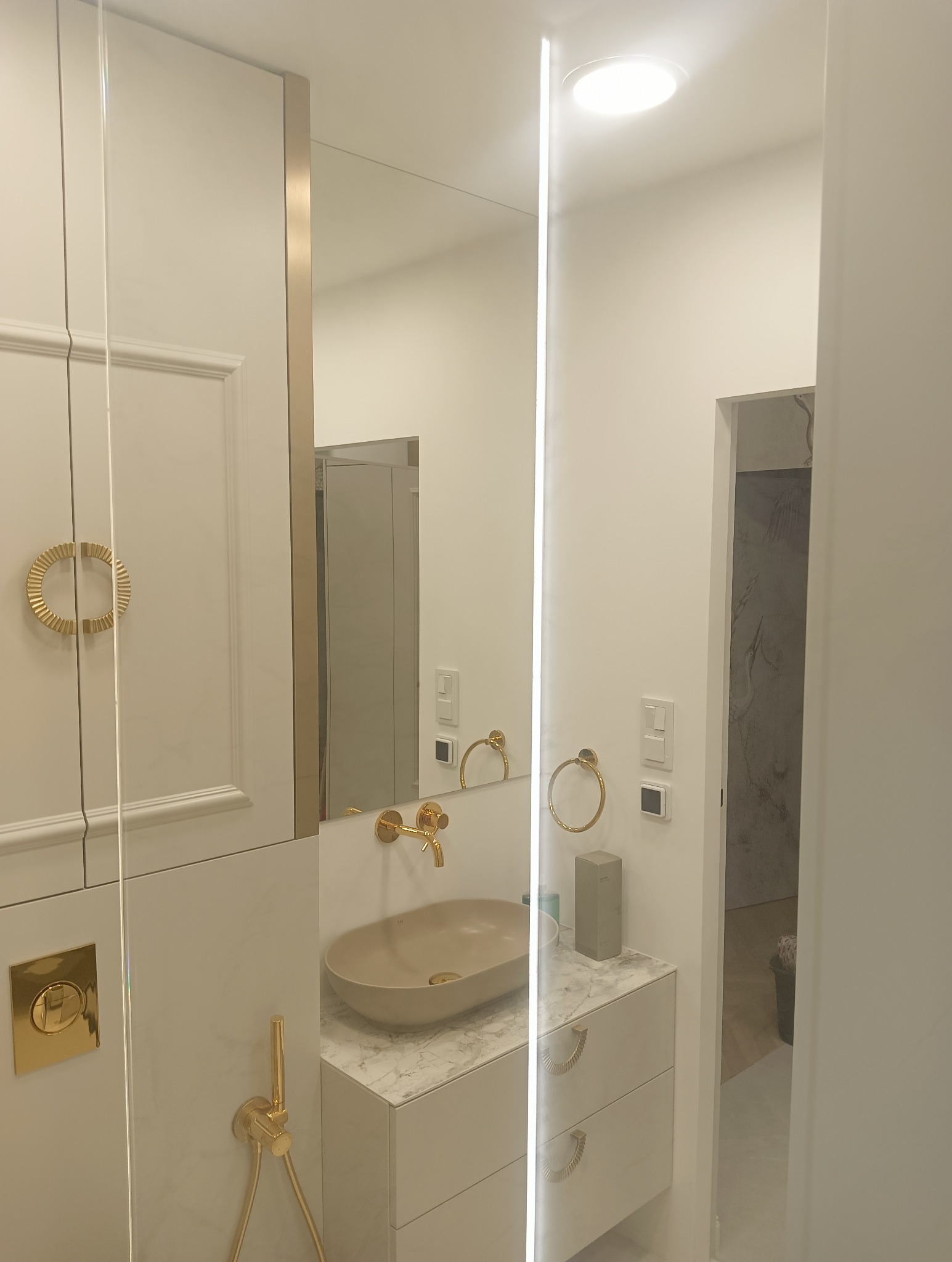 Elegancka łazienka z beżową umywalką nablatową, złotą armaturą i okrągłymi uchwytami, oświetlenie LED w lustrze, widok na fragment pomieszczenia z dekoracyjną ścianą w tle.