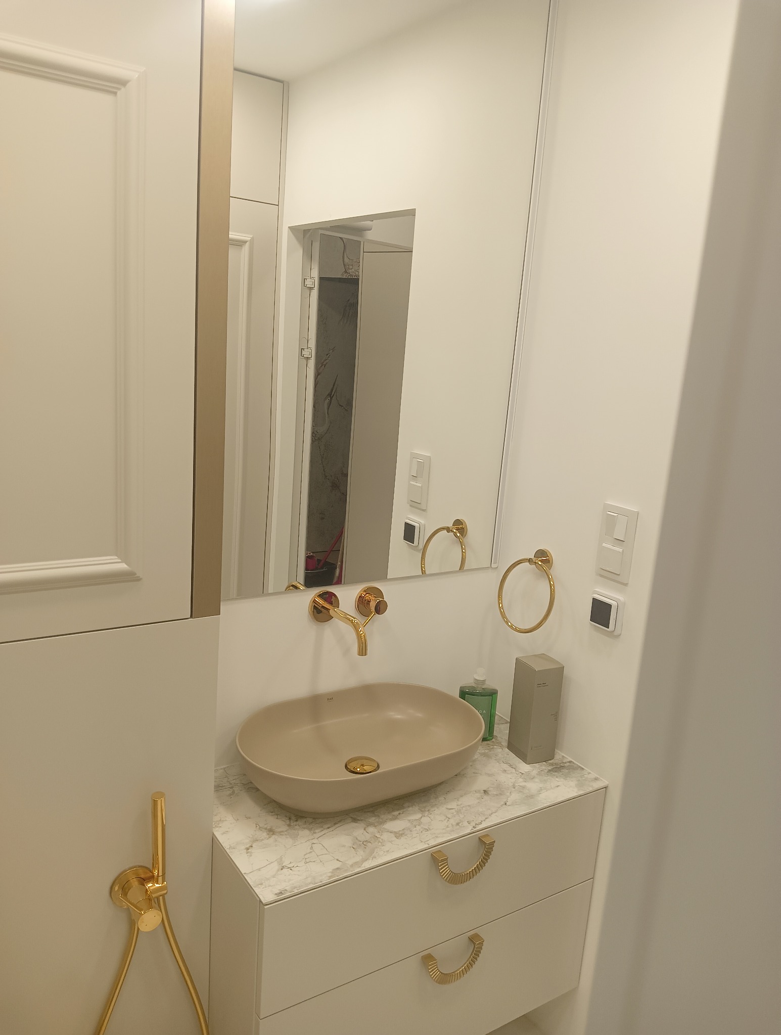 Nowoczesna łazienka z beżową umywalką nablatową i armaturą w kolorze złota, białe szafki z marmurowym blatem, akcesoria łazienkowe i widok na remontowane wnętrze w lustrze.