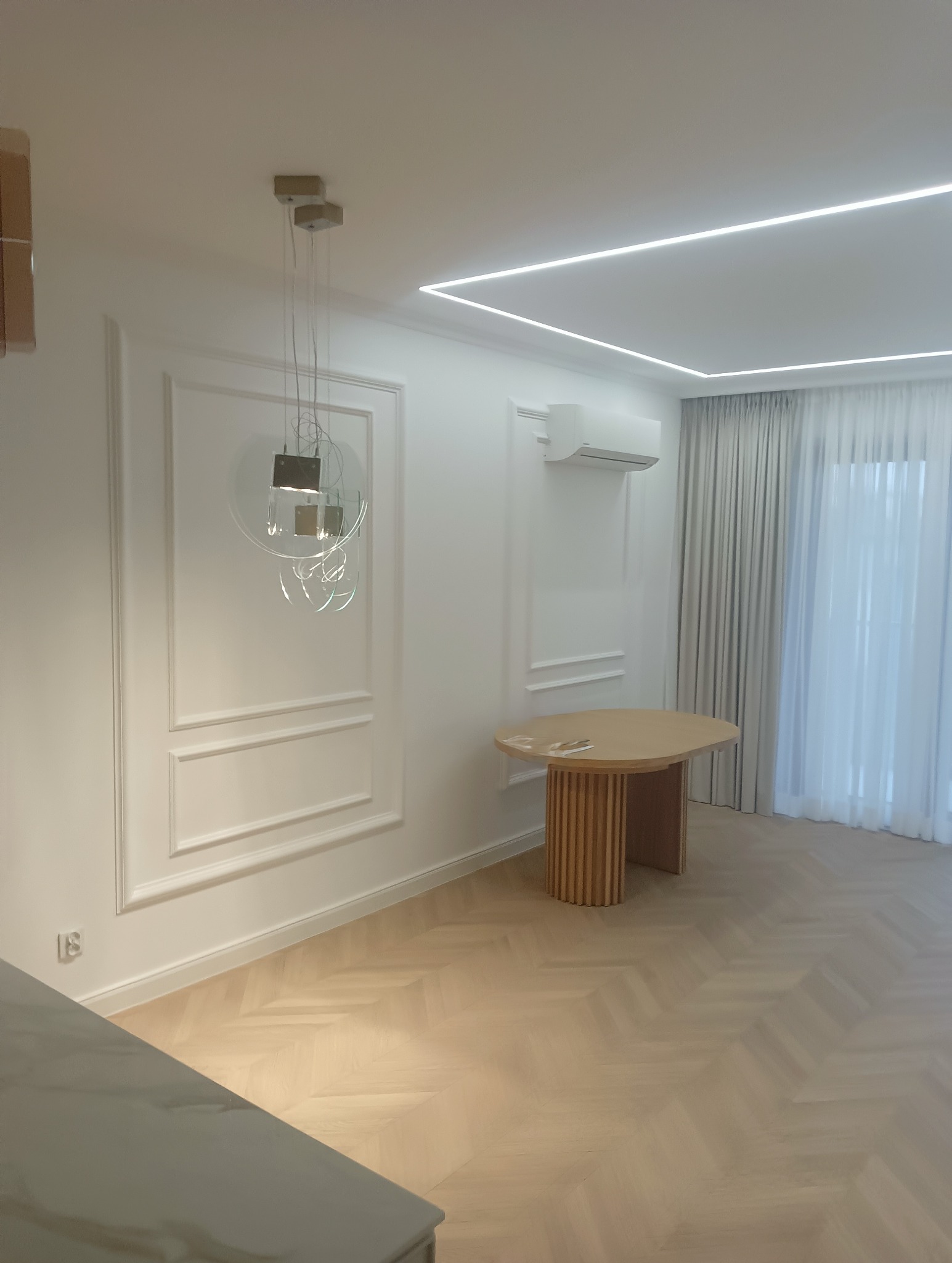 Jasne panele ułożone we wzór jodełki francuskiej, okrągły stół z żłobioną podstawą, białe ściany z dekoracyjnymi listwami i nowoczesną lampą wiszącą, klimatyzator na ścianie, białe zasłony...