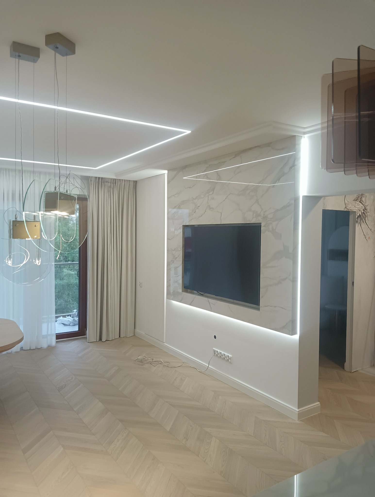 Nowoczesny salon z marmurową ścianą telewizyjną podświetloną LED, parkiet ułożony w jodełkę, designerskie lampy wiszące i zasłony w kolorze beżowym.