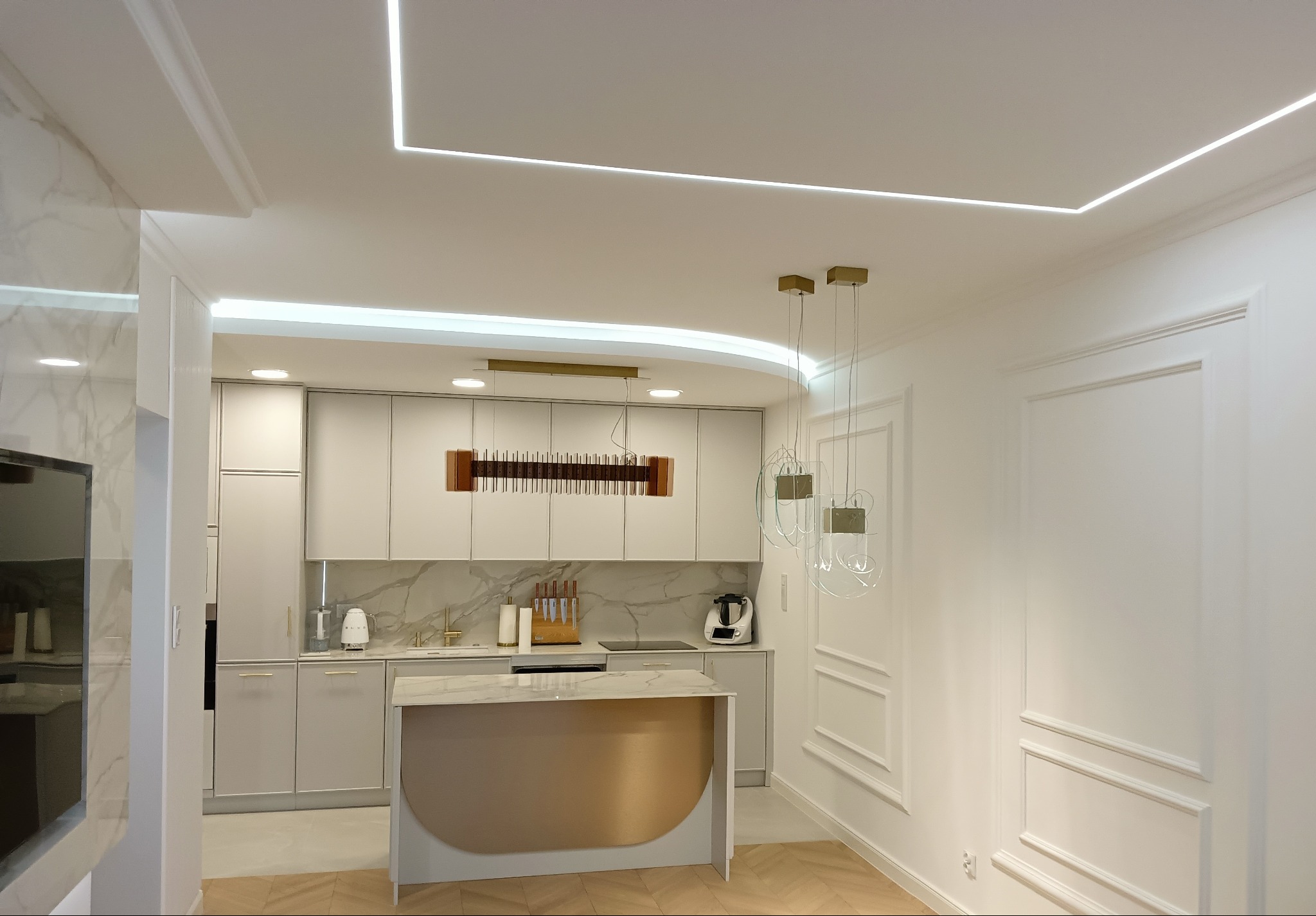 Nowoczesna kuchnia z wyspą, oświetleniem LED w suficie i dekoracyjnymi sztukateriami na ścianach, marmurowy backsplash, złote akcenty.