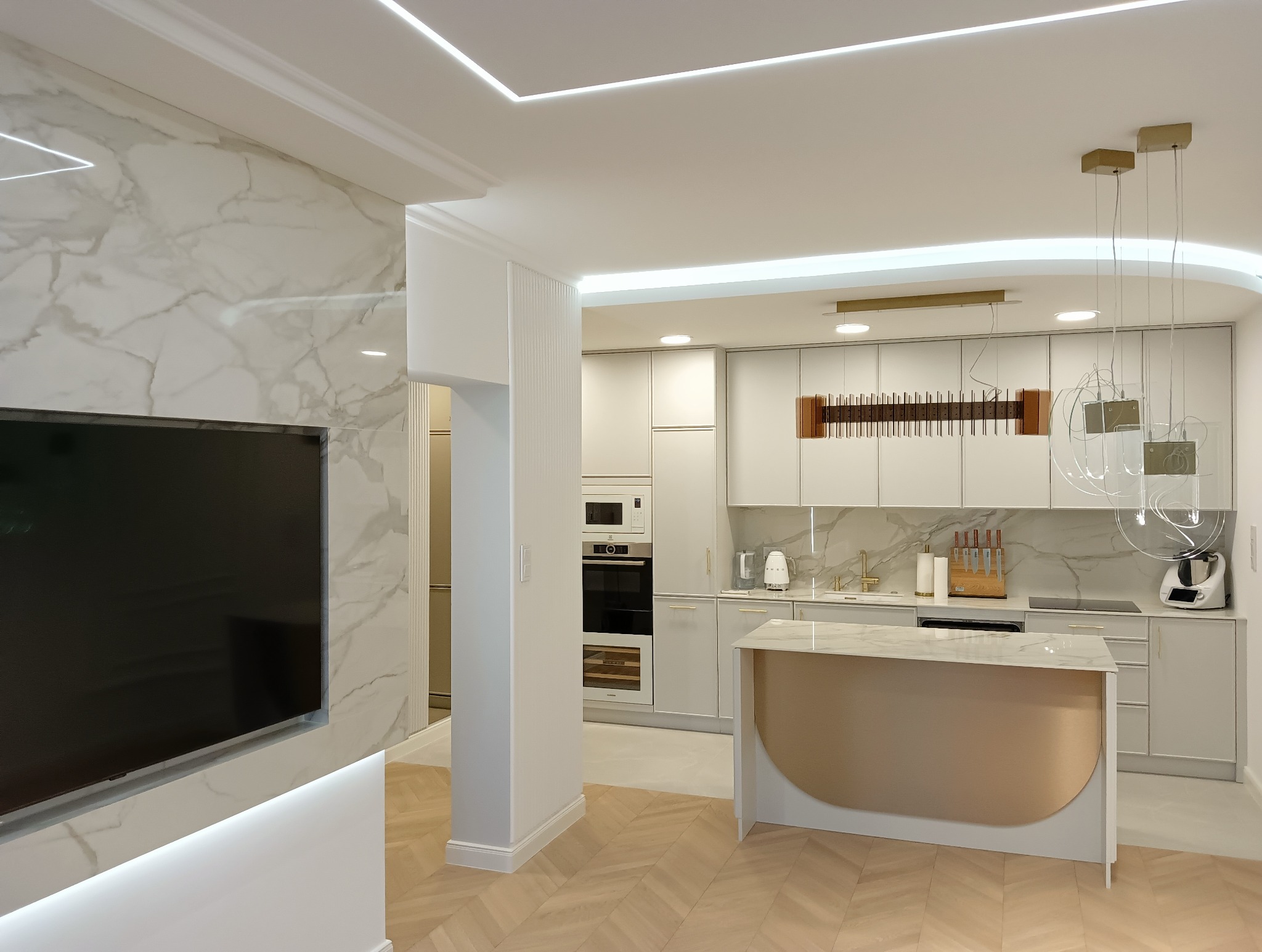 Nowoczesna kuchnia z marmurową ścianą telewizyjną, blatem i wyspą kuchenną w odcieniach bieli i złota, podświetlona listwami LED, z parkietem ułożonym w jodełkę.