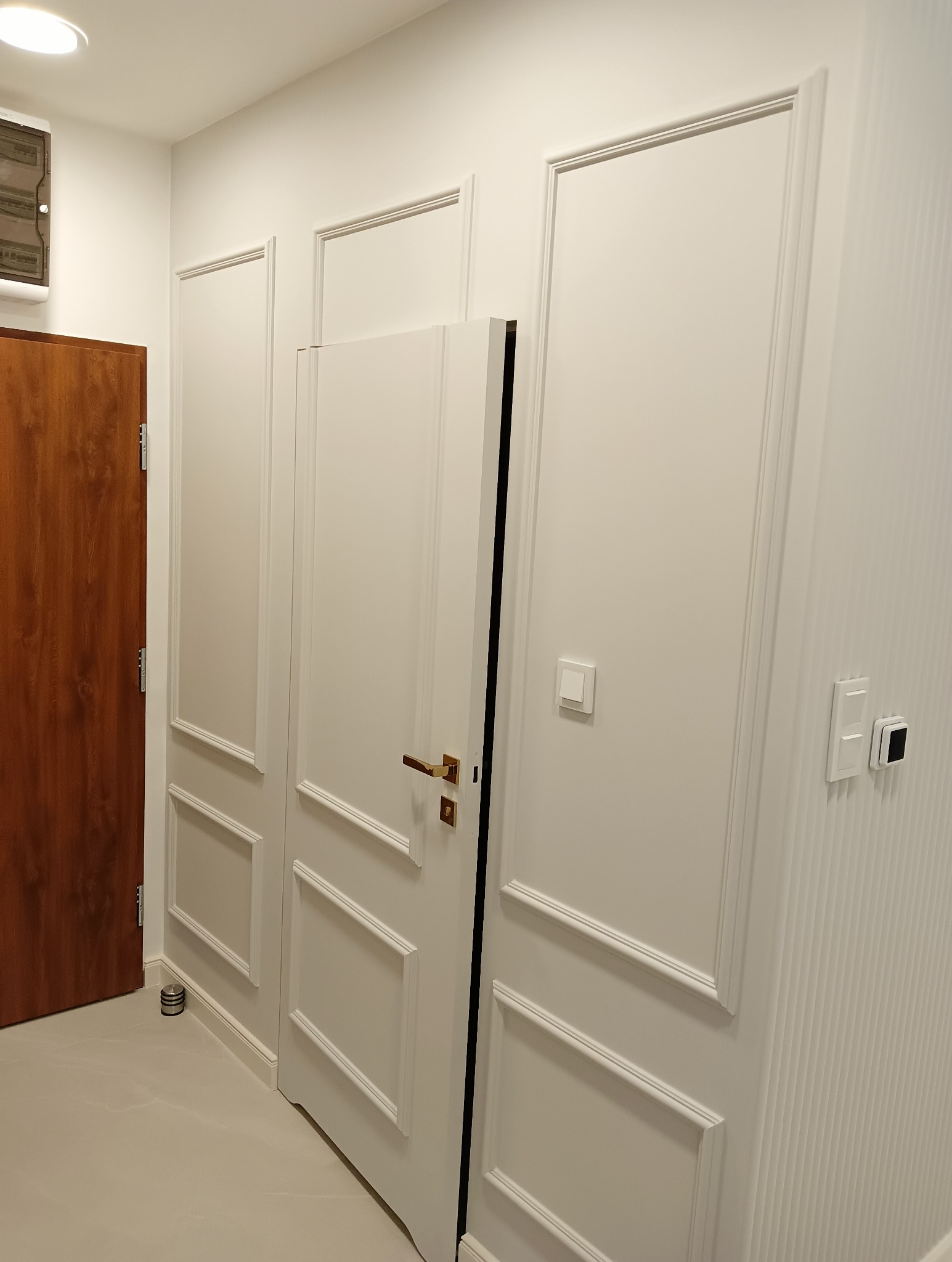 Białe drzwi wewnętrzne z ozdobnymi listwami na ścianie, widoczny brązowy element drzwi z lewej strony, szara podłoga i włączniki światła.