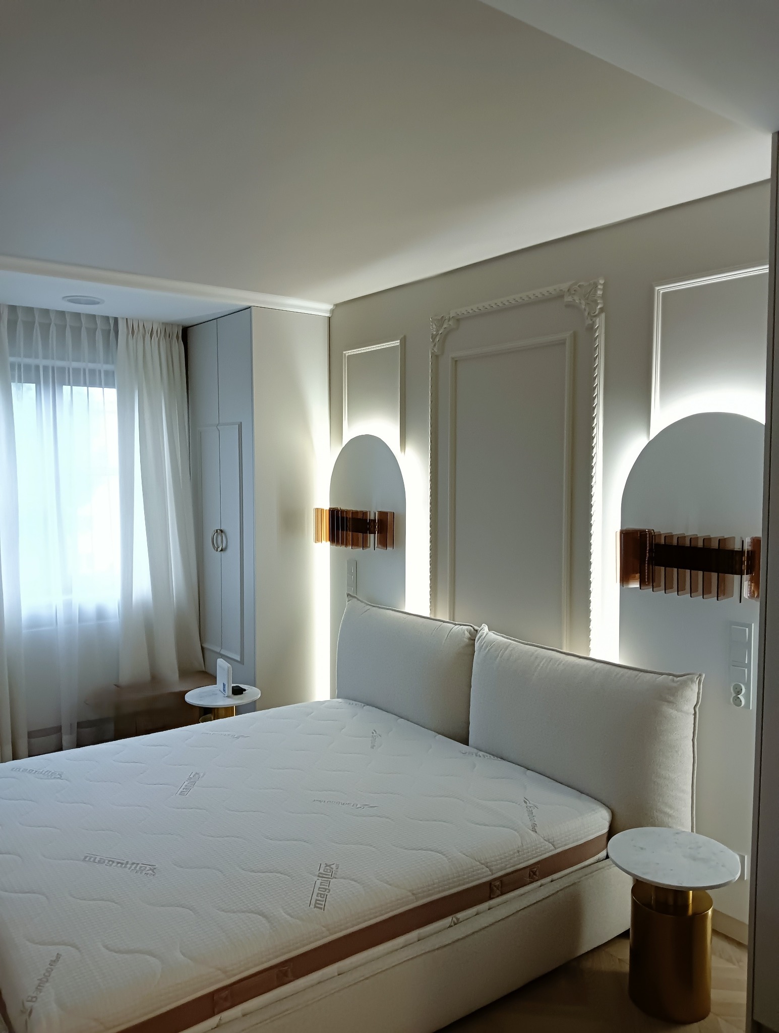 Elegancka sypialnia w jasnych barwach z łóżkiem Magniflex, oświetleniem ściennym w stylu art deco i dekoracyjnymi panelami ściennymi.