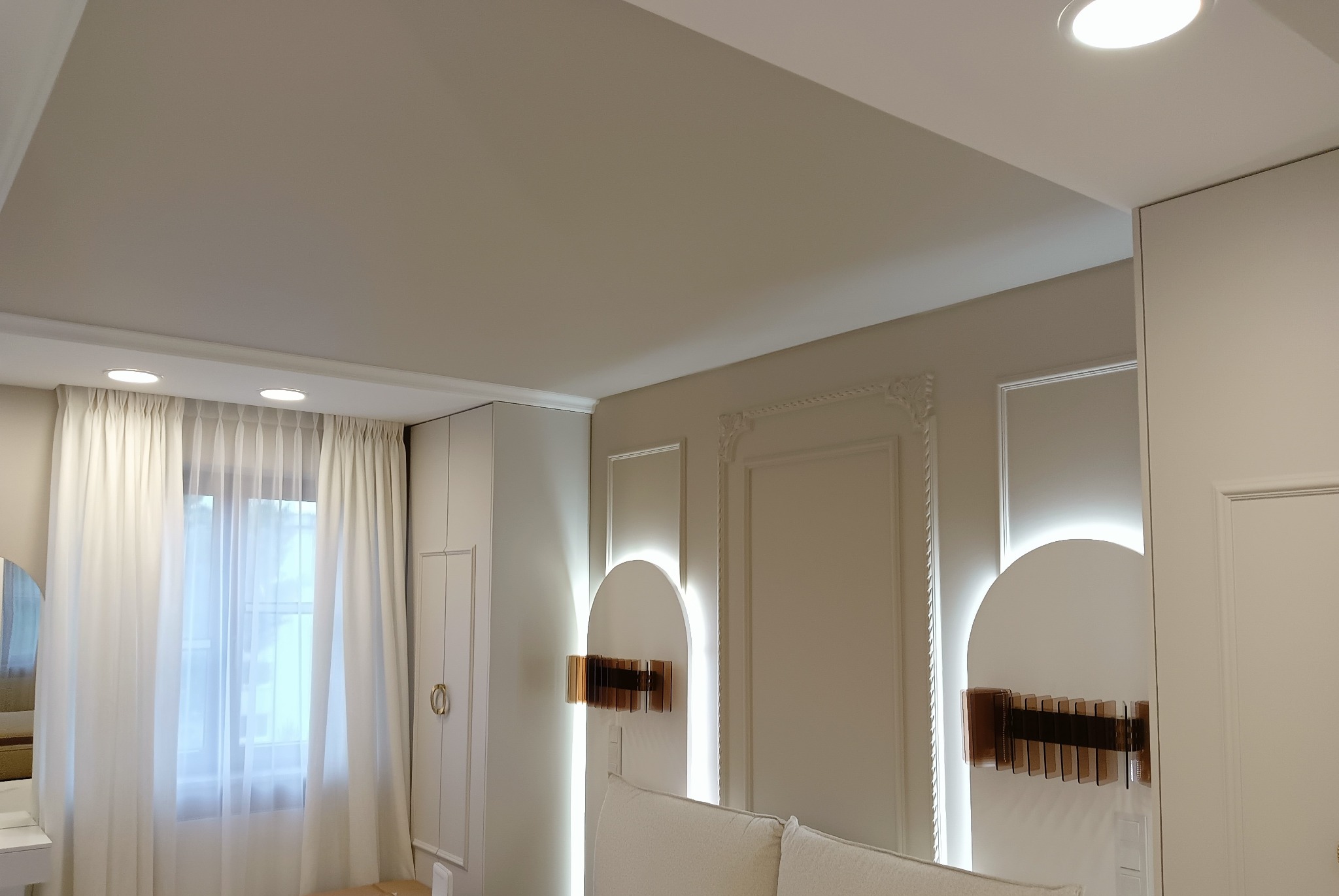 Elegancka aranżacja wnętrza z kremowymi ścianami, sztukaterią, wbudowanymi lampami LED, białymi zasłonami i nowoczesnymi dekoracjami ściennymi z podświetleniem.