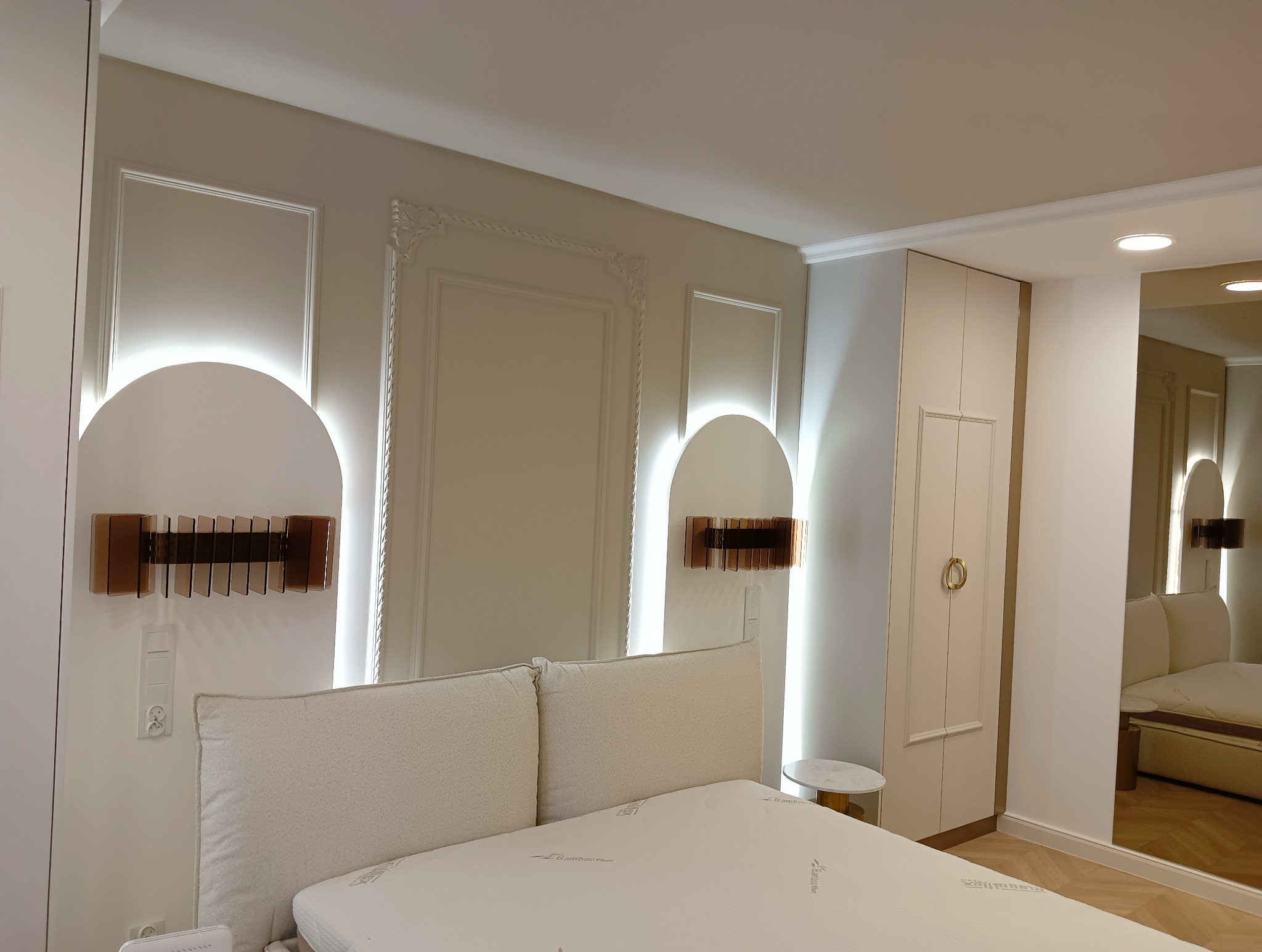 Elegancka sypialnia z beżowymi ścianami, dekoracyjnymi listwami, podświetlanymi wnękami i nowoczesnymi lampkami nocnymi z brązowymi akcentami, widok na łóżko z kremowym zagłówkiem i szafę...