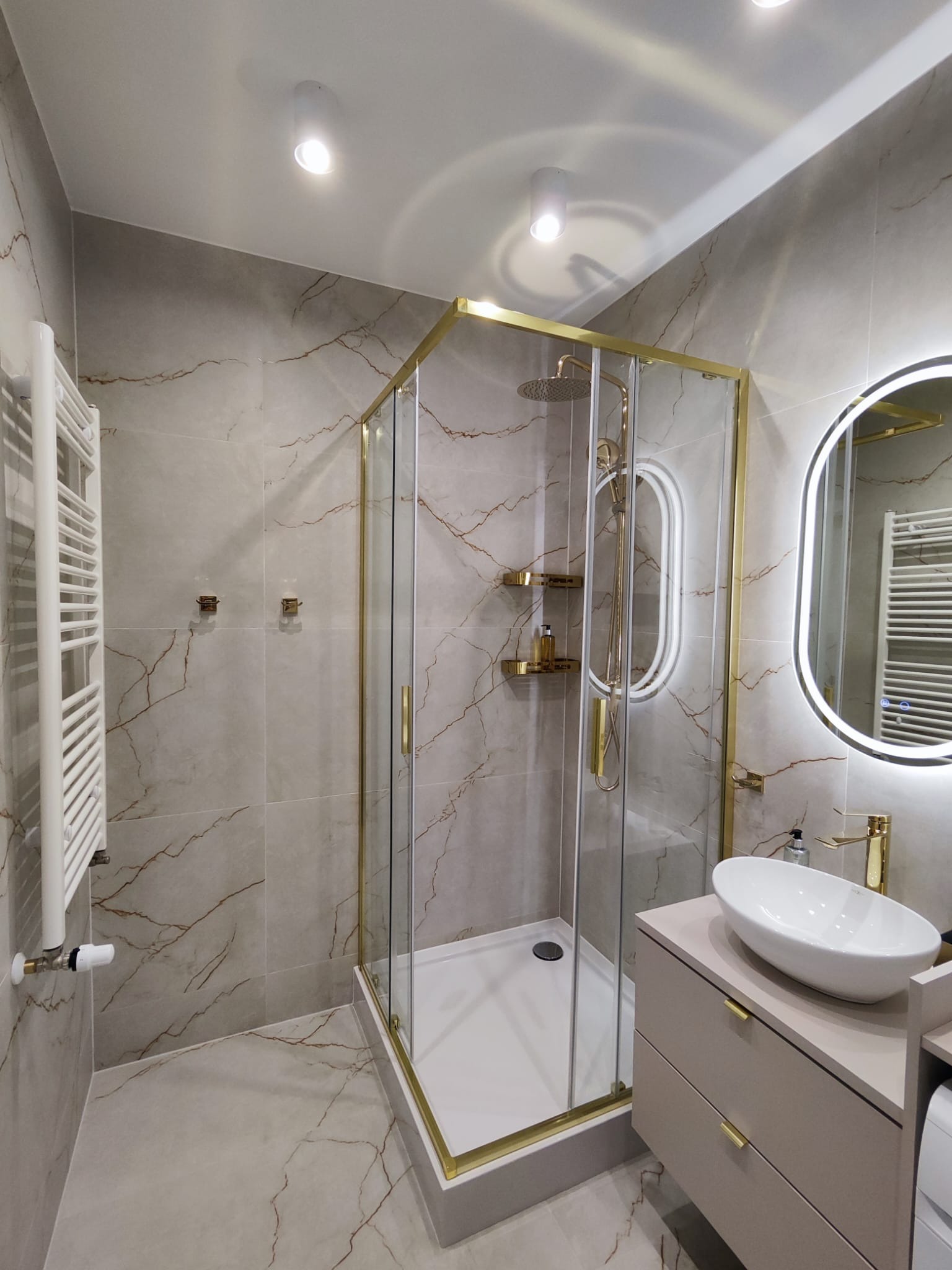 Elegancka łazienka w szarościach z akcentami złota: kabina prysznicowa z złotymi ramami, umywalka nablatowa i podświetlane lustro owalne.