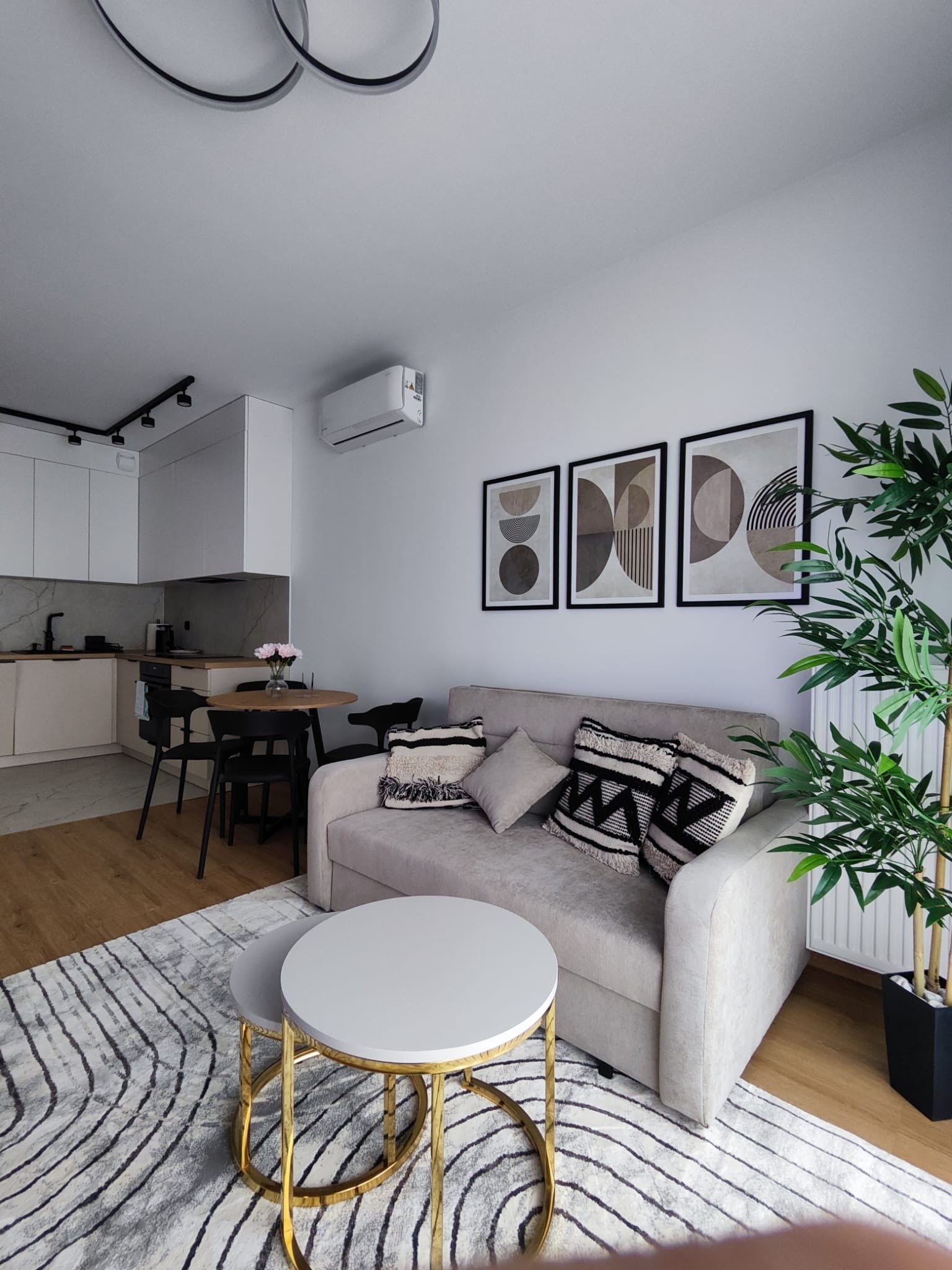 Jasne wnętrze salonu z aneksem kuchennym. Beżowa sofa z poduszkami, okrągły stolik kawowy z złotymi nogami, dywan w geometryczne wzory. Na ścianie grafiki w ramkach.