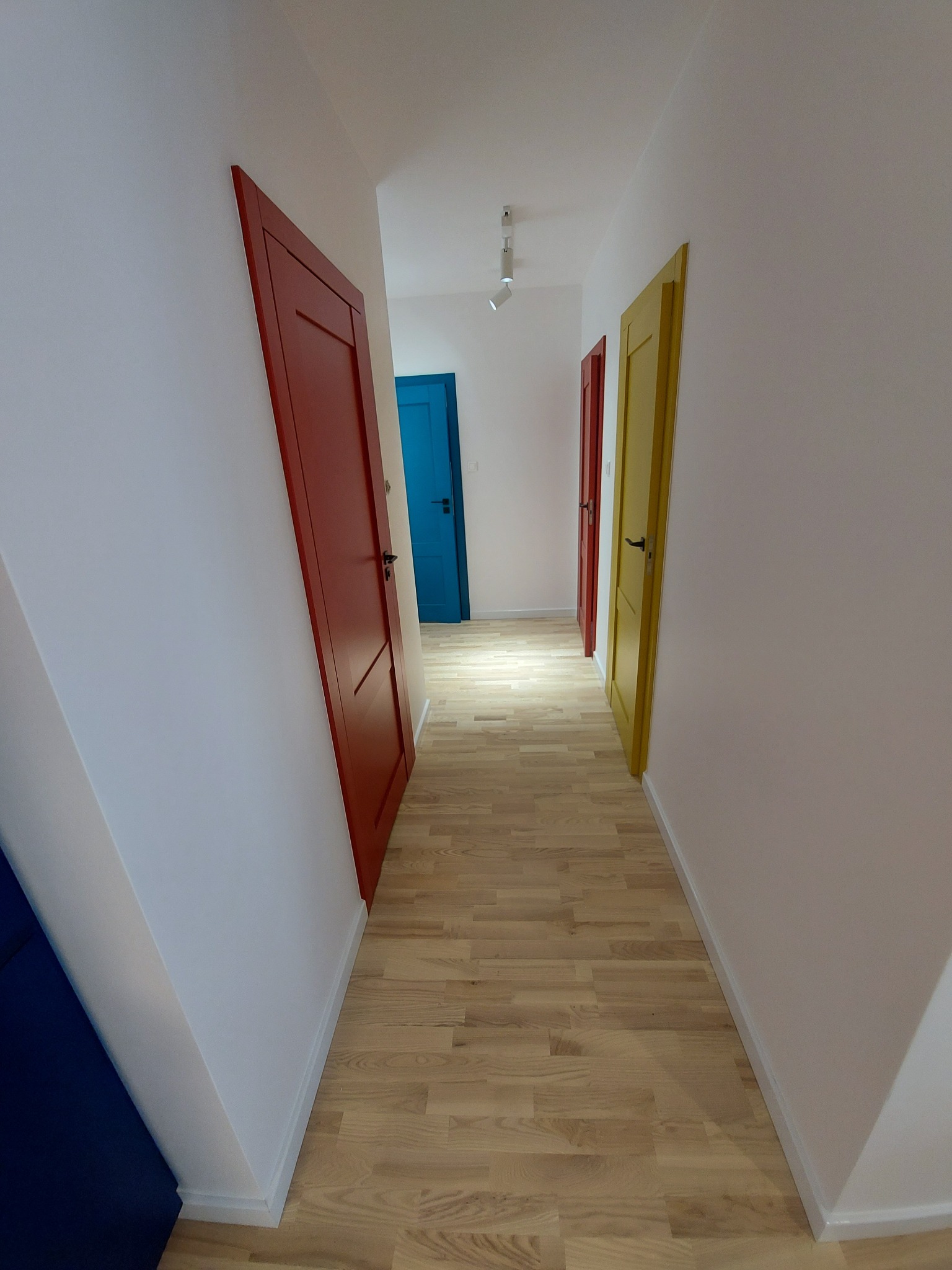 Korytarz po remoncie z parkietem i kolorowymi drzwiami (czerwone, żółte, niebieskie). Białe ściany, oświetlenie sufitowe punktowe. Perspektywa z głębi korytarza.