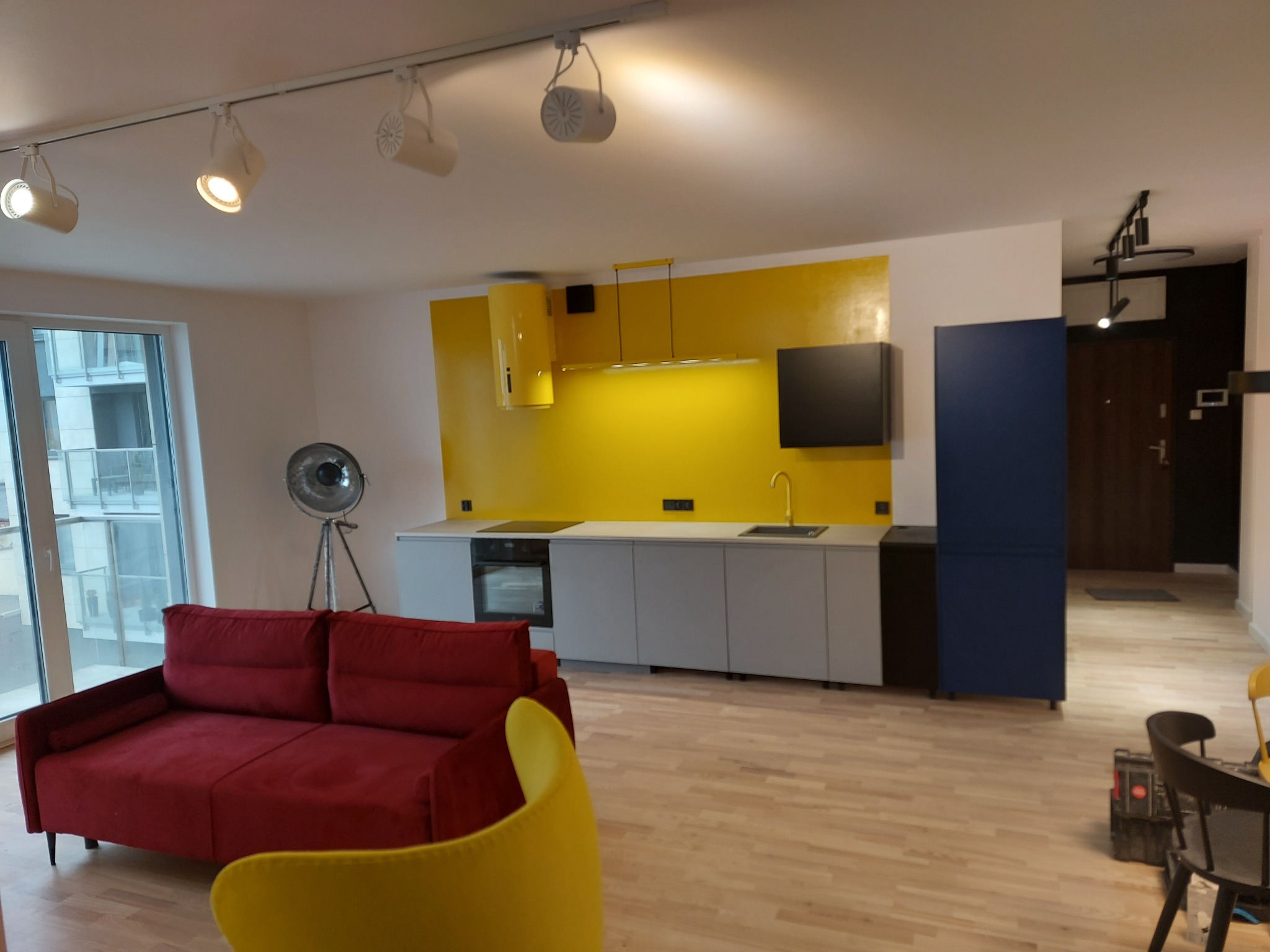 Żółto-szara kuchnia z czerwonym narożnikiem i parkietem. Nowoczesne oświetlenie sufitowe, okno z widokiem na miasto. Elementy dekoracyjne w stylu industrialnym.