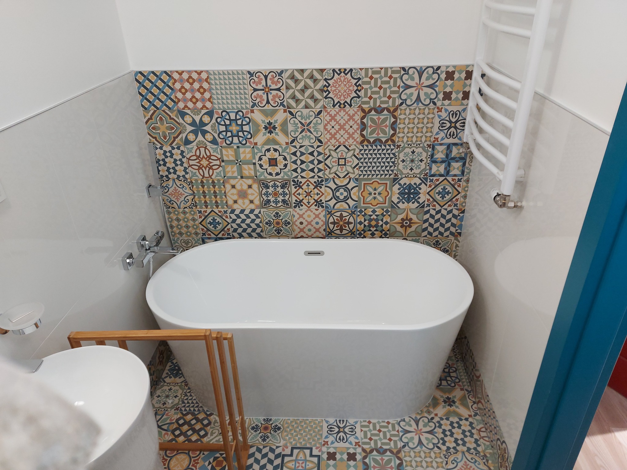 Biała wanna wolnostojąca na tle ściany z mozaiką w łazience. Widoczny również grzejnik drabinkowy i umywalka z drewnianym stelażem.