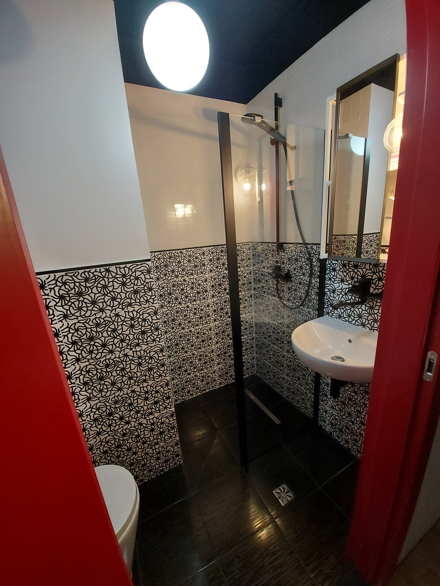 Nowoczesna łazienka z kabiną prysznicową, umywalką i czarno-białymi wzorzystymi płytkami. Czerwona rama drzwi dodaje kontrastu. Widoczne oświetlenie sufitowe.
