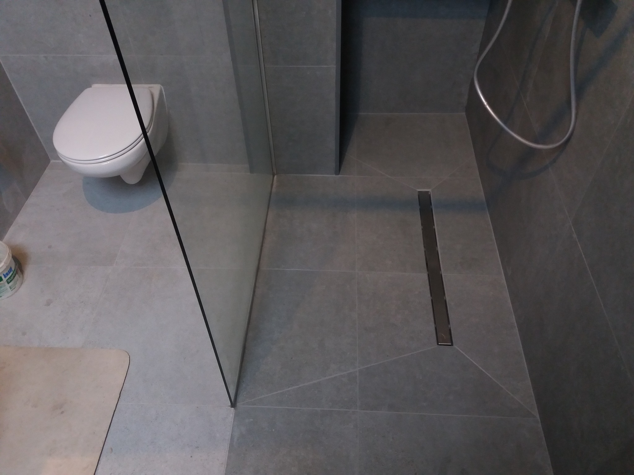 Nowoczesna łazienka z szarymi płytkami, kabiną prysznicową typu walk-in i odpływem liniowym. Widoczny fragment toalety podwieszanej i węża prysznicowego.