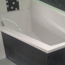 360 GLOBAL INVEST SPÓŁKA Z OGRANICZONĄ ODPOWIEDZIALNOŚCIĄ - Biała wanna obudowana ciemnymi, strukturalnymi płytkami, z dekoracyjnym pasem płytek mozaikowych i oświetleniem LED w niszy ściennej.