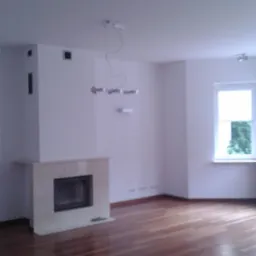 Jasne wnętrze pokoju z parkietem, widocznym kominkiem z beżowego kamienia i nowoczesną lampą wiszącą. Świeżo wykończone, puste pomieszczenie z oknem.