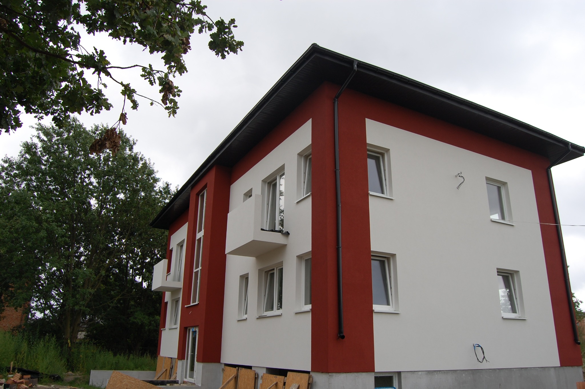 Dwukondygnacyjny dom murowany z białymi ścianami i czerwonymi elementami, z małymi balkonami, widok z dołu na elewację frontową z widocznymi przewodami elektrycznymi wychodzącymi ze ściany.