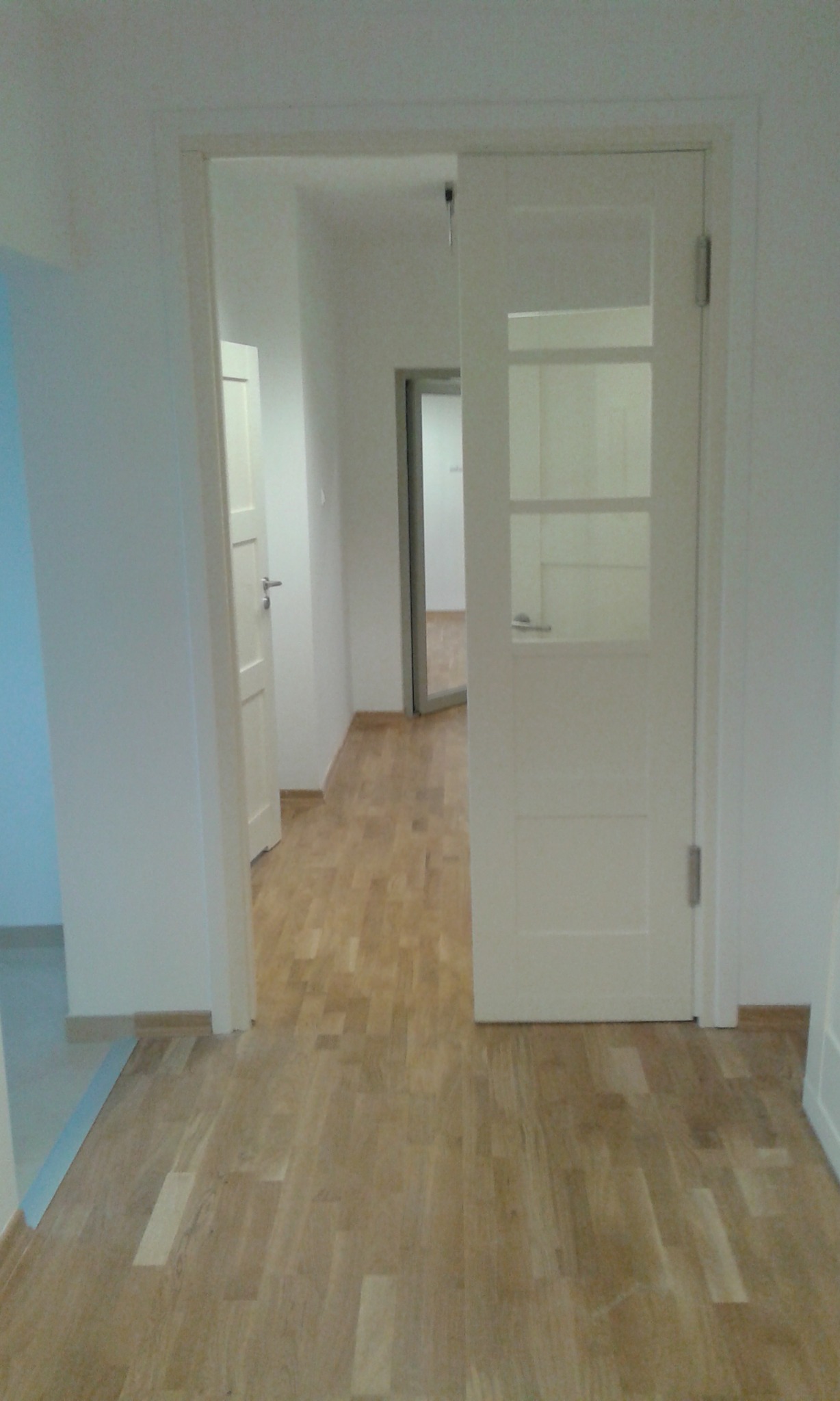 Jasne, drewniane panele podłogowe w nowym mieszkaniu, widok na białe drzwi z przeszkleniami i otwarte drzwi w głębi, świeżo pomalowane ściany.