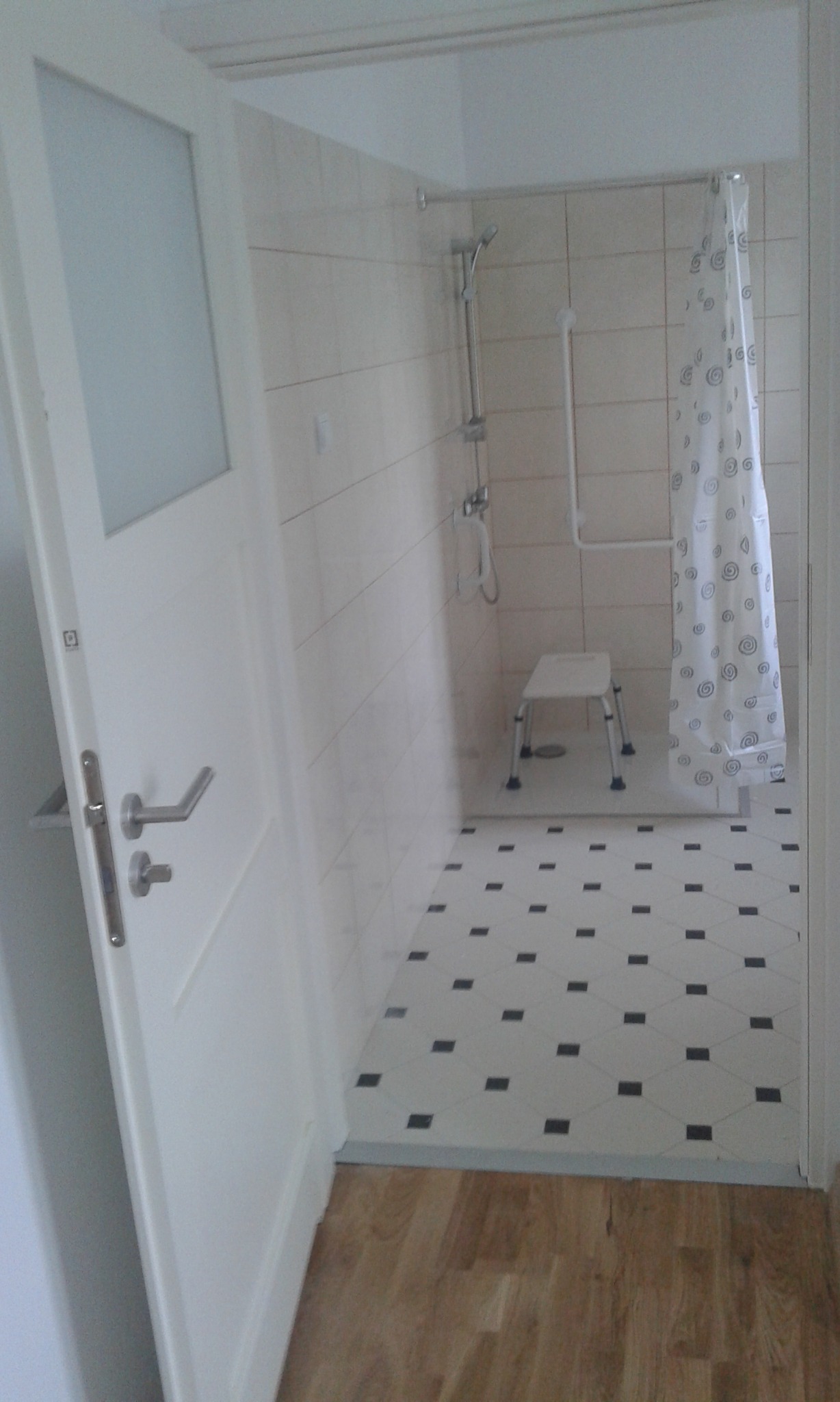 Jasna łazienka z kabiną prysznicową, białą zasłoną w szare wzory, chromowaną armaturą i uchwytem, oraz siedziskiem dla osoby niepełnosprawnej; podłoga wyłożona biało-czarną mozaiką.
