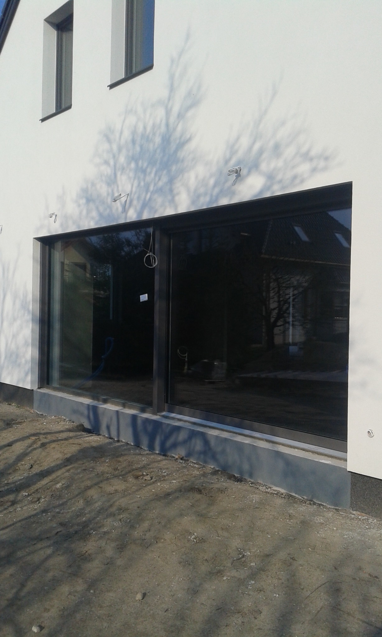 Duże, przesuwne okno w surowym stanie na tle białej ściany budynku, widoczne cienie drzew i elementy instalacji na elewacji.