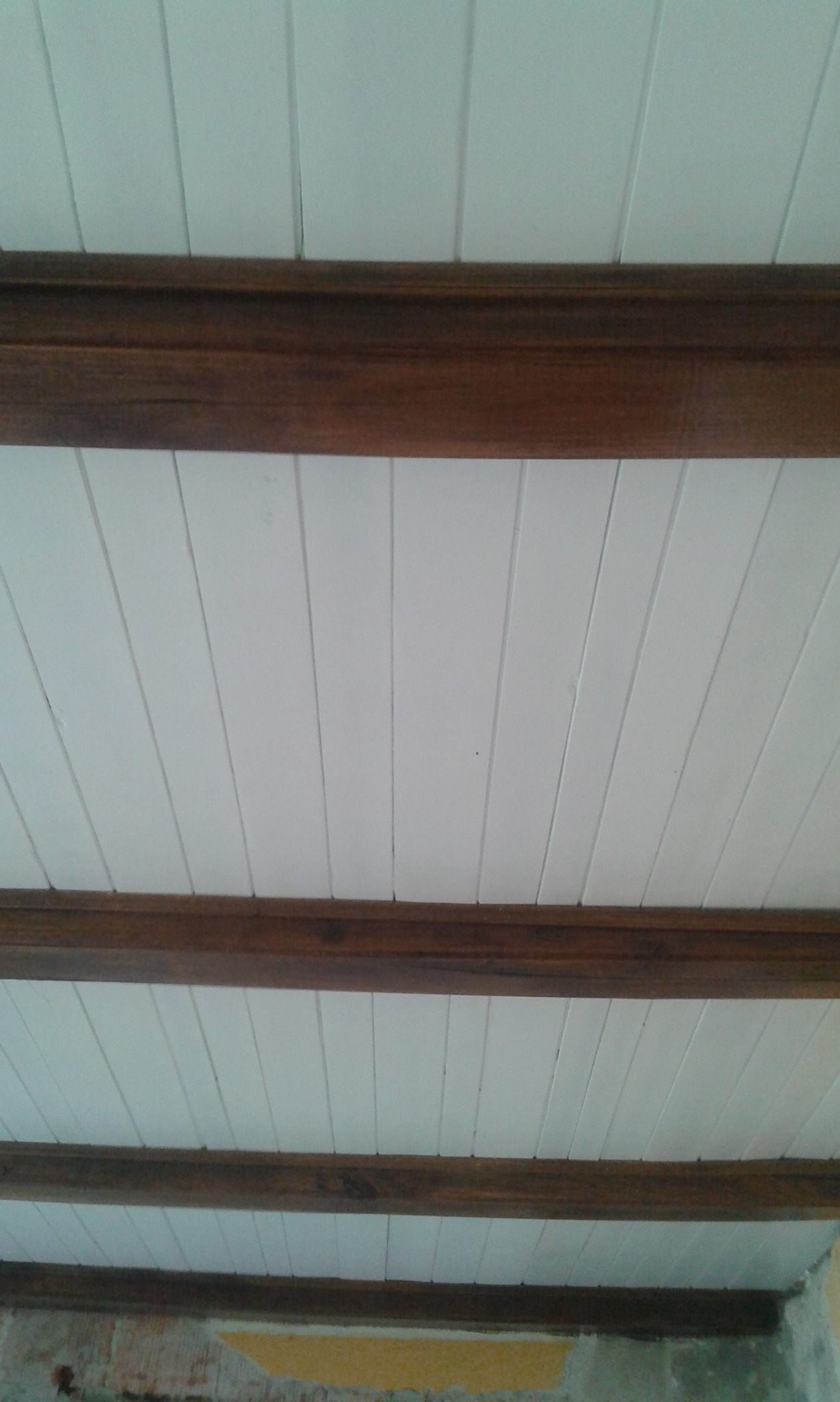 Biały sufit z drewnianymi belkami, widoczne ślady remontu na ścianie w tle.