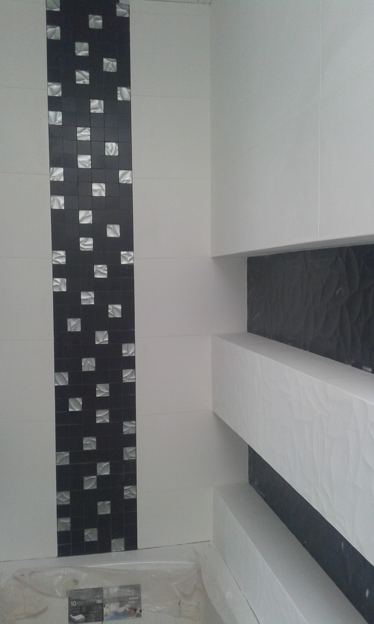 Białe płytki ścienne z czarnym, pionowym pasem mozaiki z kwadratowymi elementami i geometryczne półki z czarnym tłem, częściowo zakryte folią ochronną na podłodze.