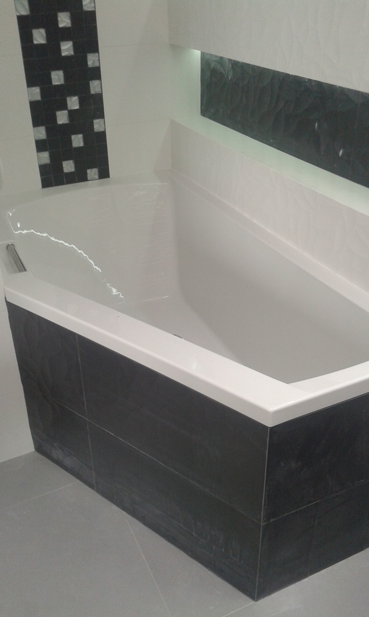 Biała wanna obudowana ciemnymi, strukturalnymi płytkami, z dekoracyjnym pasem płytek mozaikowych i oświetleniem LED w niszy ściennej.